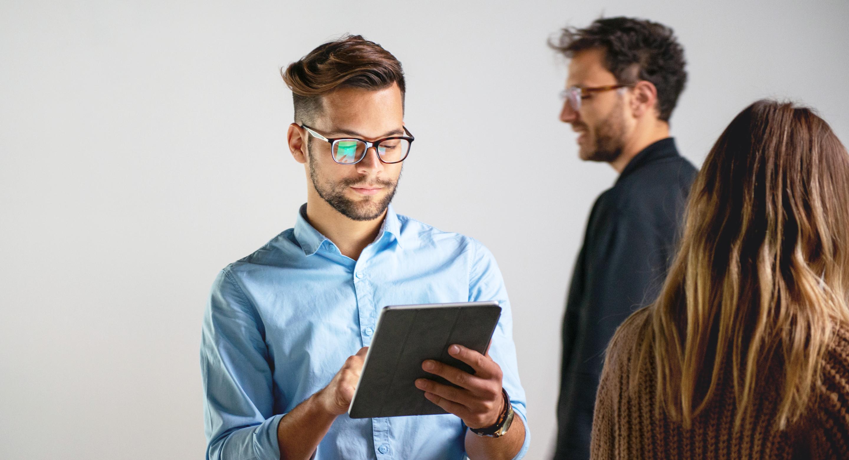 A person using a tablet while two colleagues talk in the background.