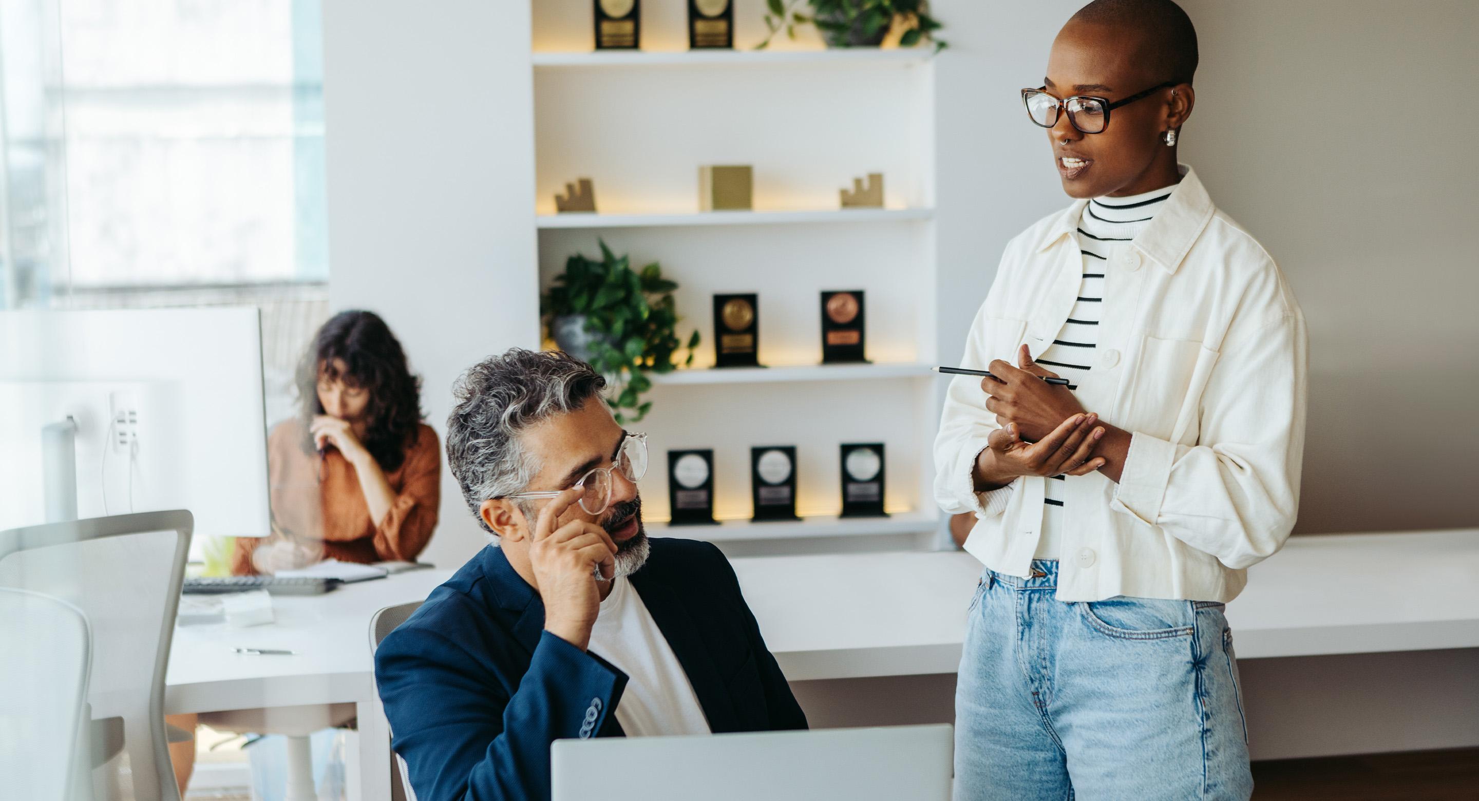 Professional woman speaking with a seated colleague in a modern office, holding a tablet.