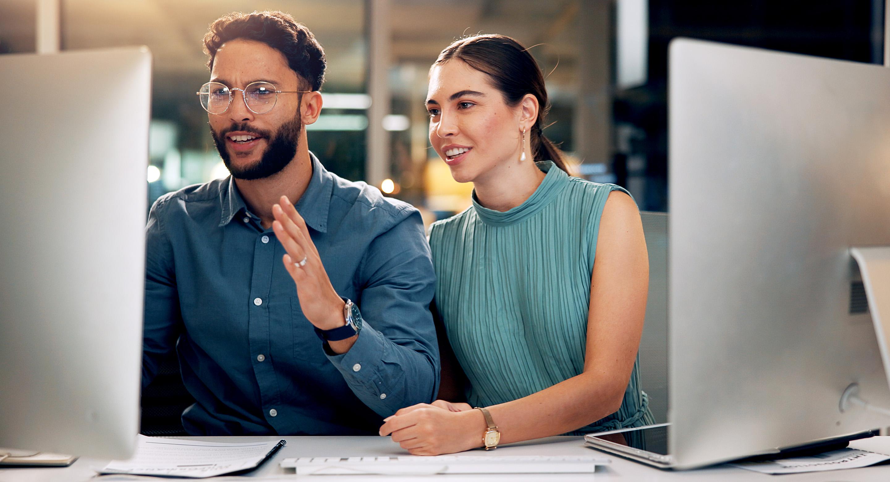 Two colleagues collaborating at desktop computers in an office, smiling while reviewing work together on screen.