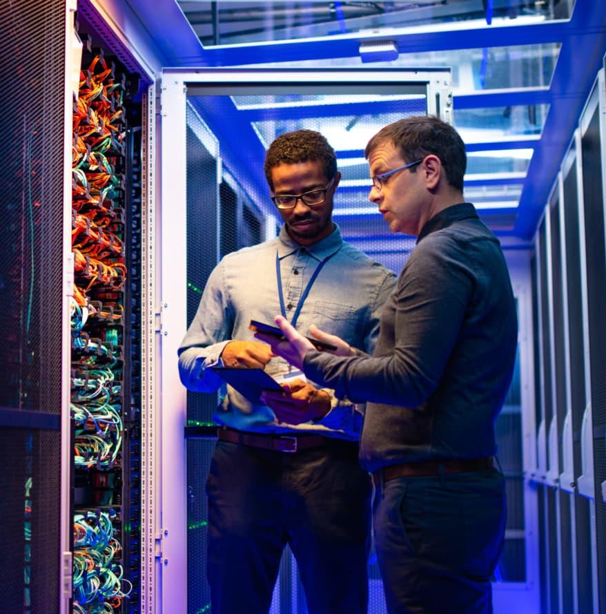 Two employees in a cable room. Both employees are carrying tablets and are in discussion.