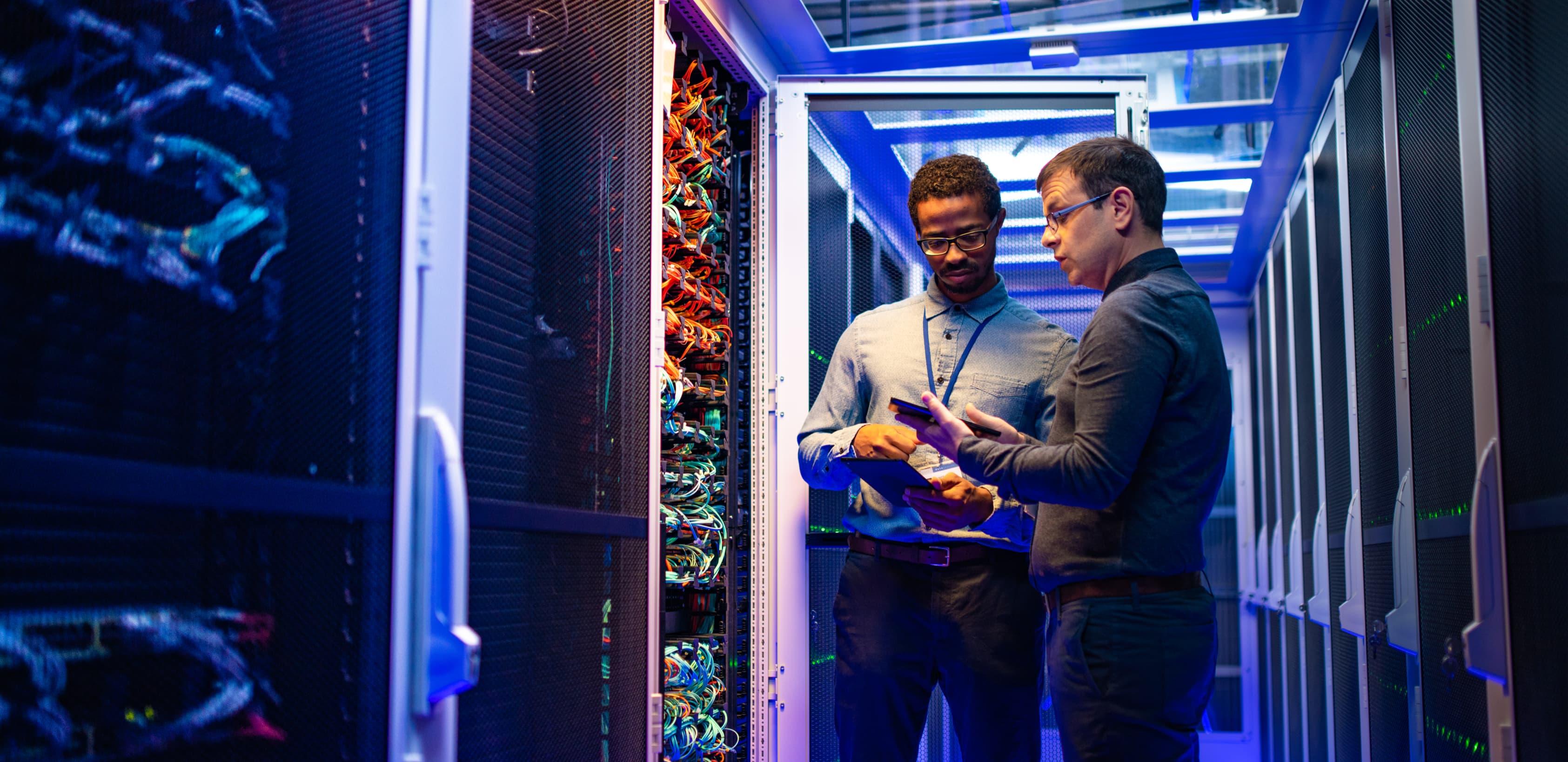 Two employees in a cable room. Both employees are carrying tablets and are in discussion.