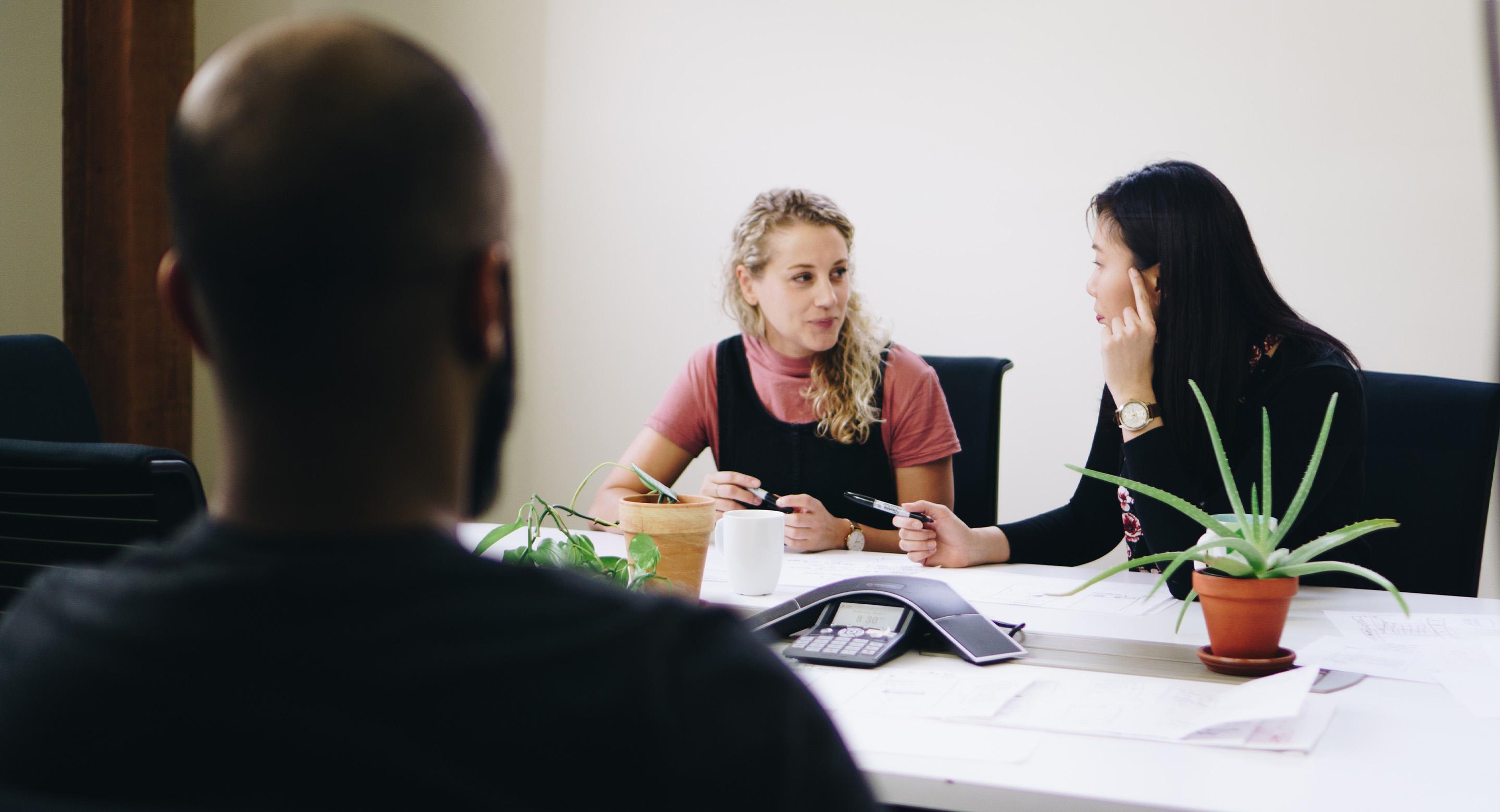 Two coworkers talking at a conference table with papers and plants, while another colleague listens from the foreground.