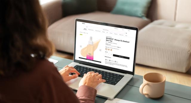 A woman browsing the IPSY website and viewing a beauty product on the laptop with a coffee cup beside it.