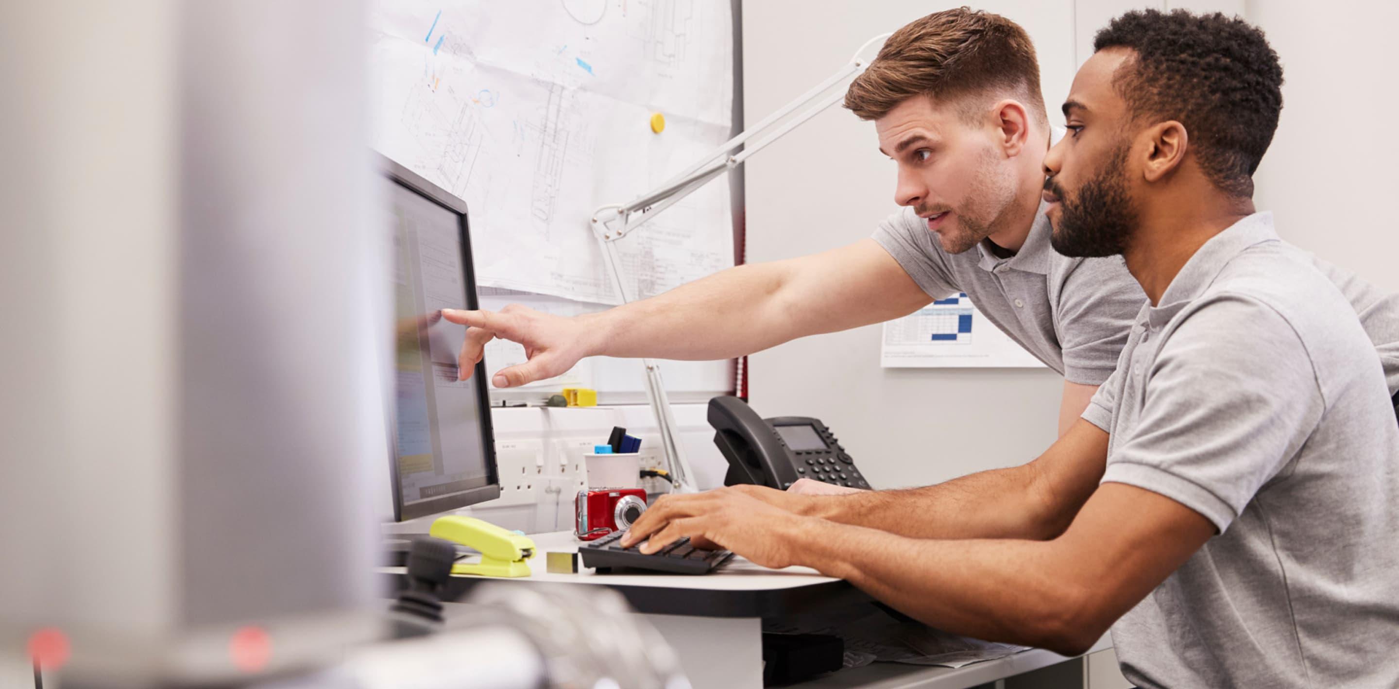 Two coworkers are working at a computer screen. One is sitting down and typing while the other one is standing up and pointing to the screen.