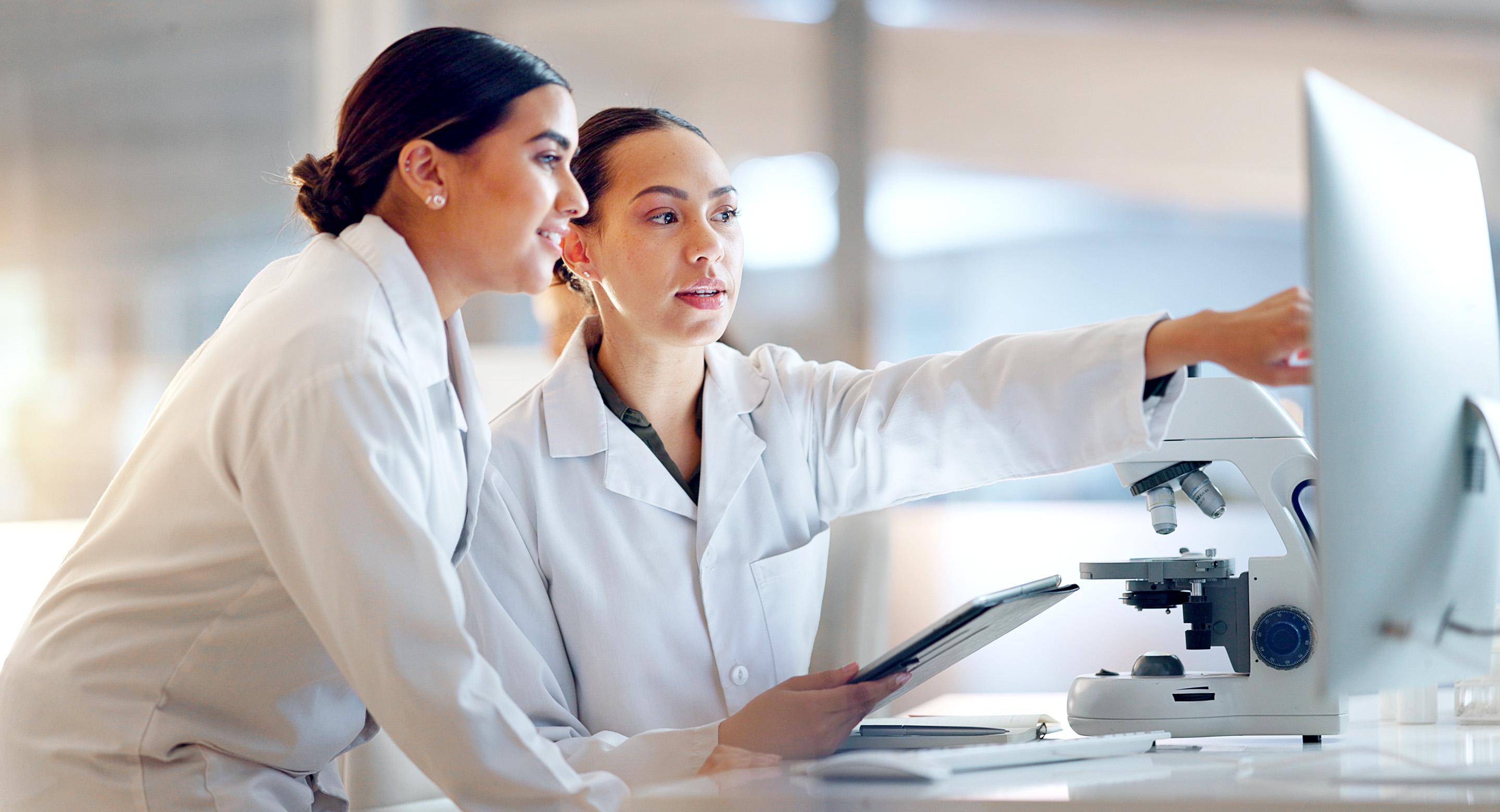 Dos trabajadores de laboratorio en batas blancas revisando información en un monitor y una tableta junto a un microscopio.