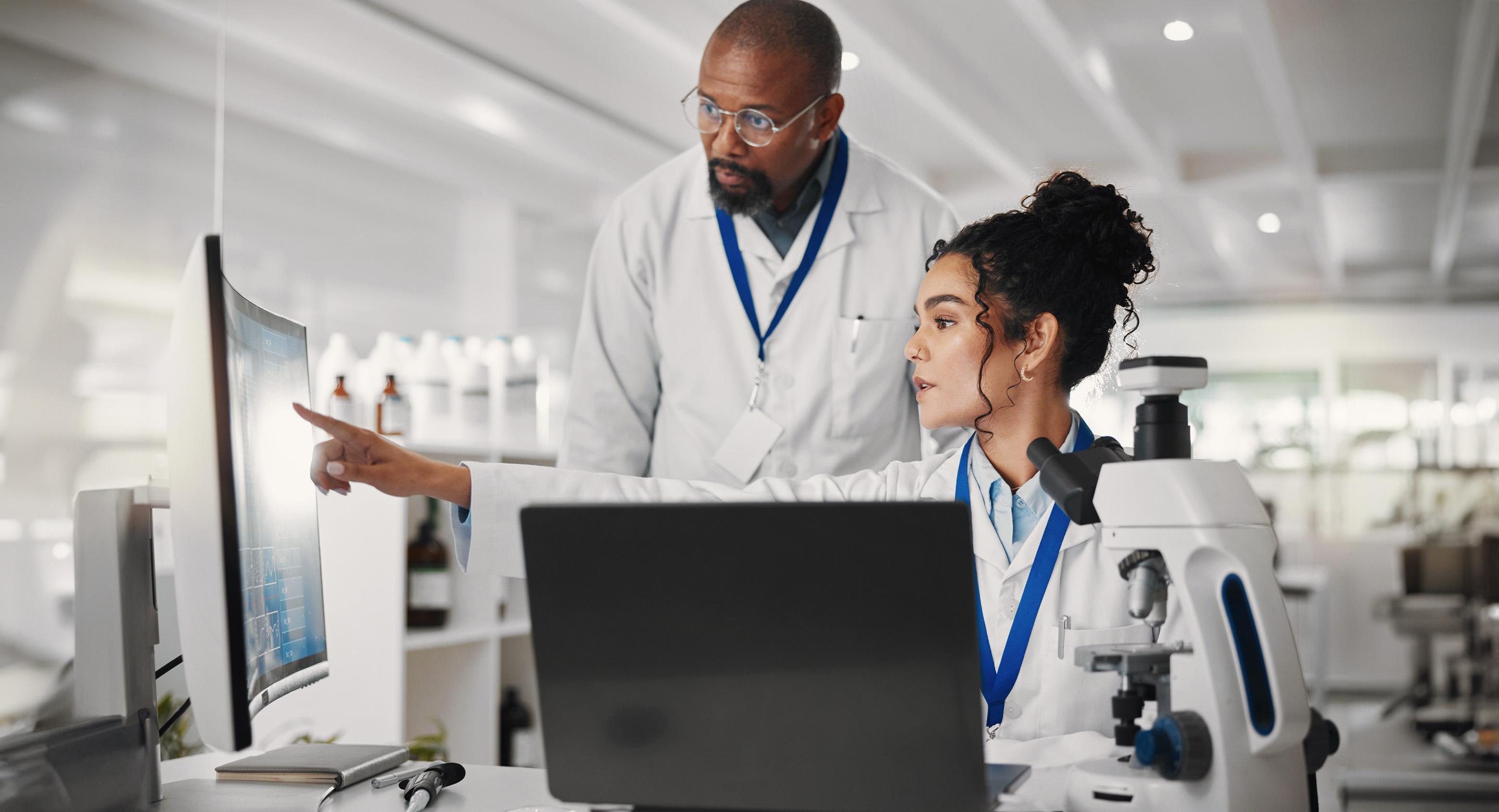 Dos trabajadores de laboratorio mirando datos en un monitor junto a un microscopio en un laboratorio.