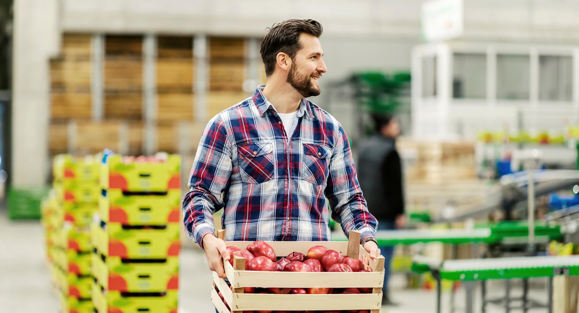 Worker in a plaid shirt carries a crate of red apples inside a wholesale food distribution warehouse.