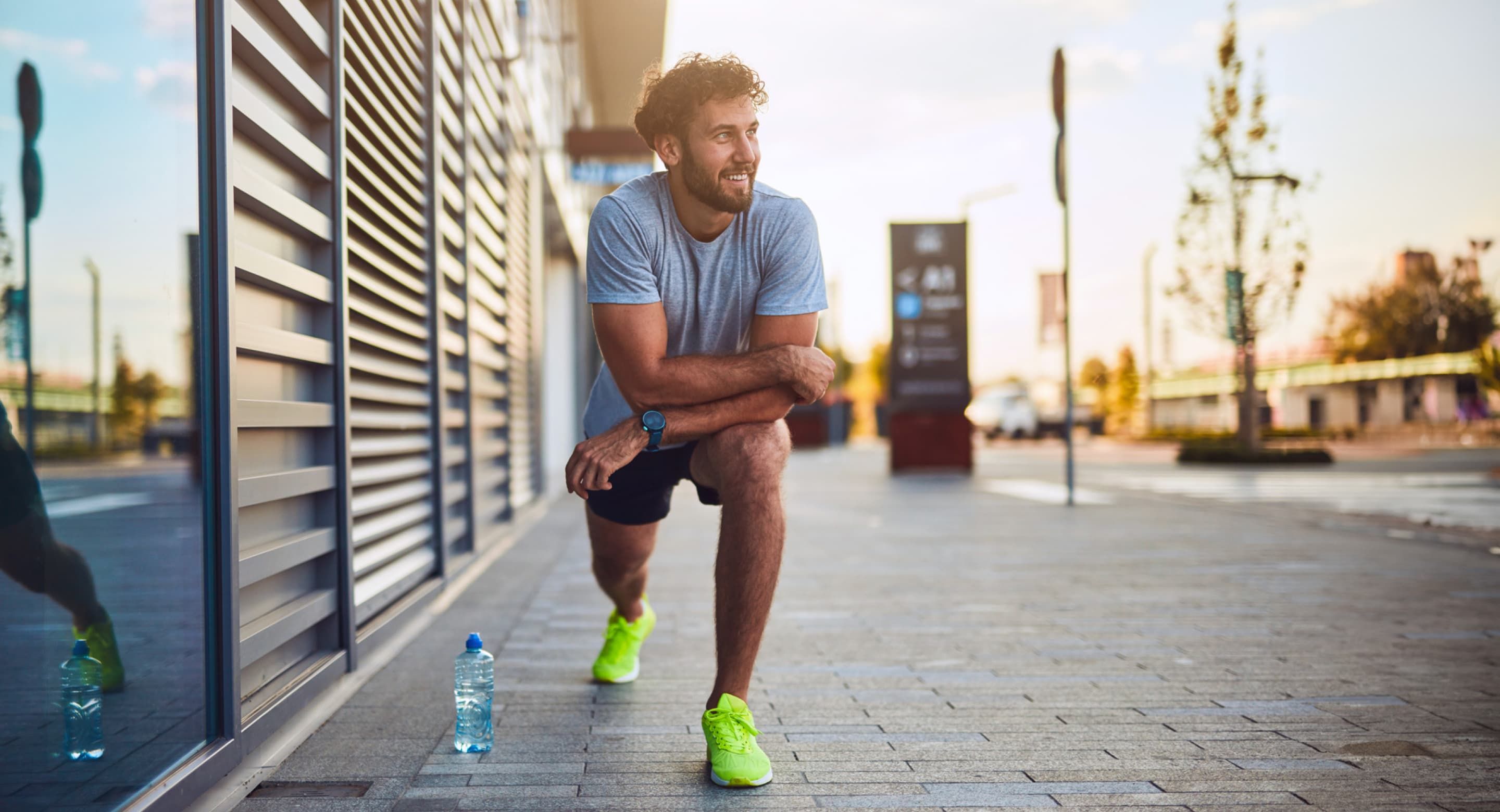 A man smiling in a lunge position on a sidewalk, wearing lime green running shoes with a small water bottle next to him.