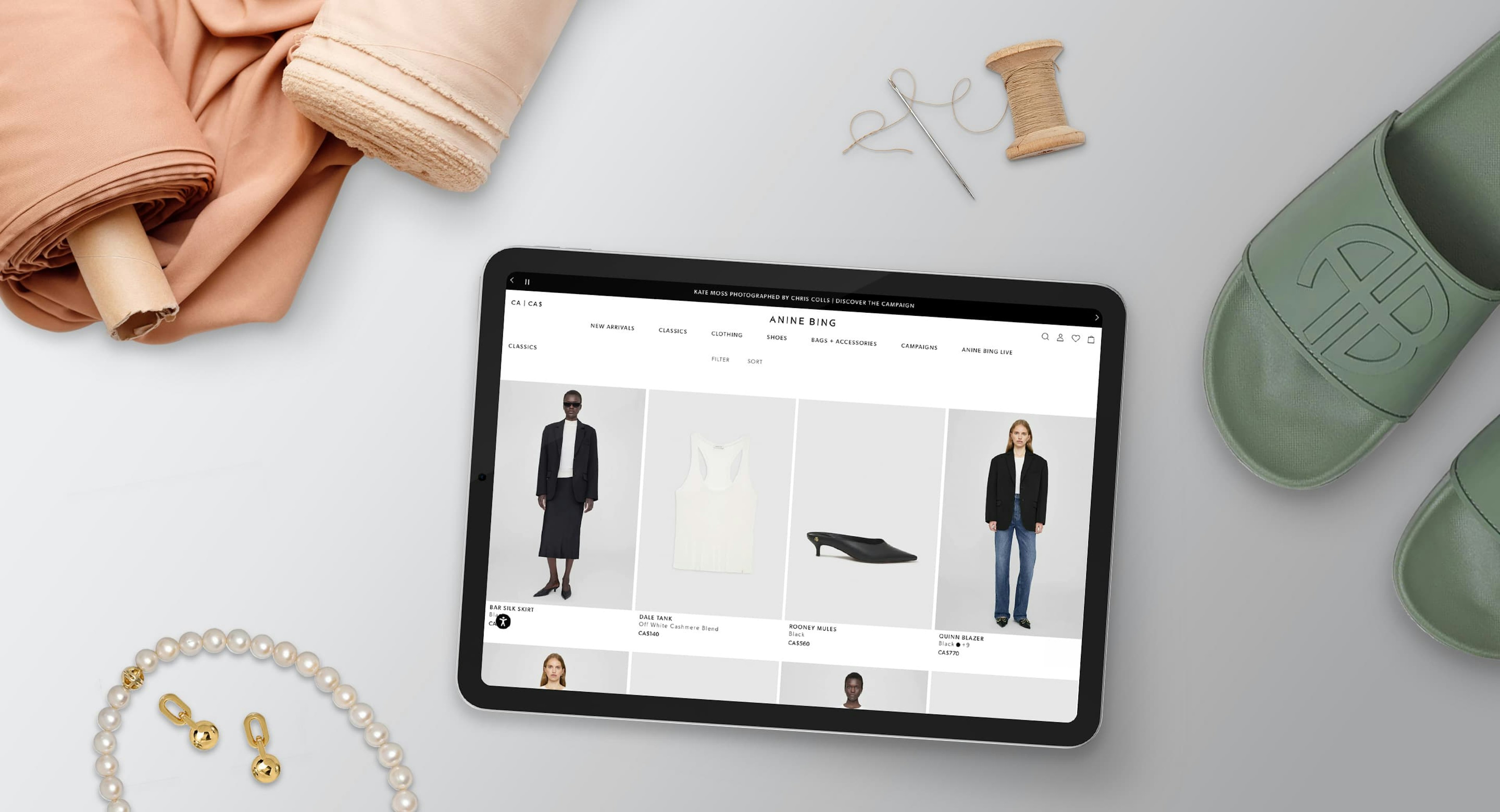 A flatlay on a grey surface including an iPad with the ANINE BING website on it. A pair of shoes, fabric rolls, a needle and thread, and a necklace and earring set surround the iPad.