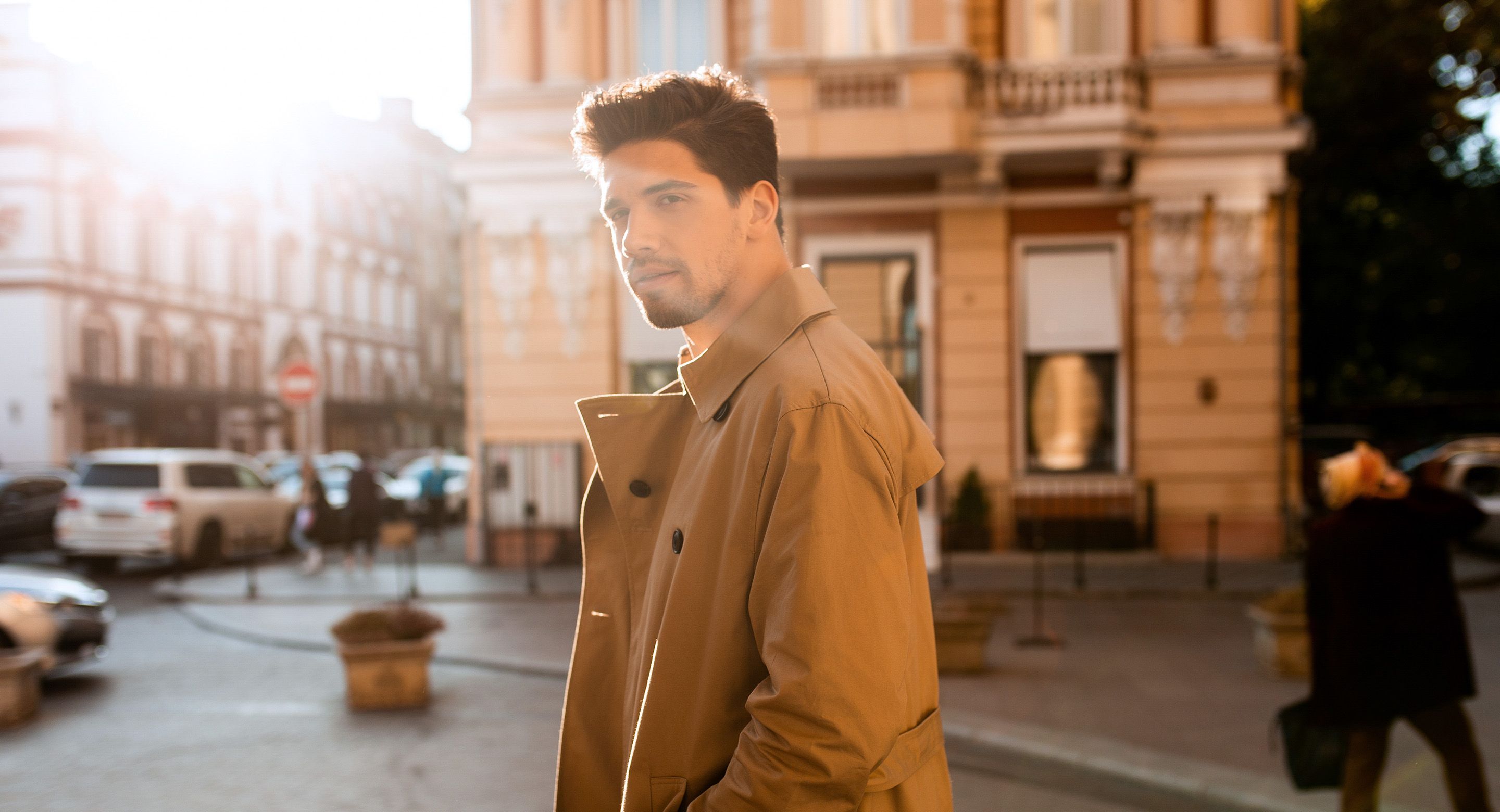 A man in a tan trench coat stands on a sunlit city street, looking toward the camera.