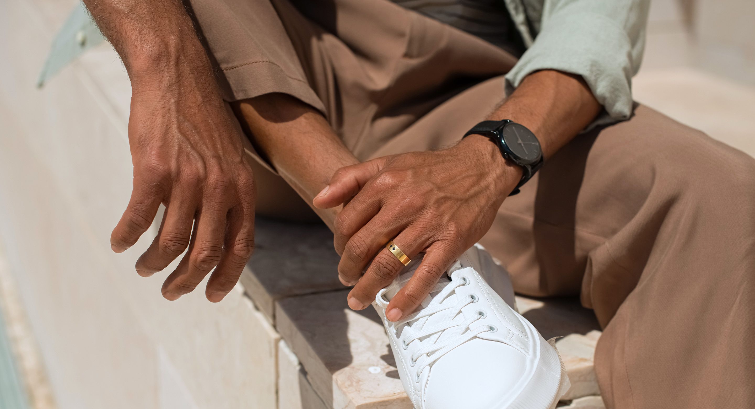 A close-up of a man’s hands, wearing a ring and watch, tying white sneakers while sitting on a stone surface.