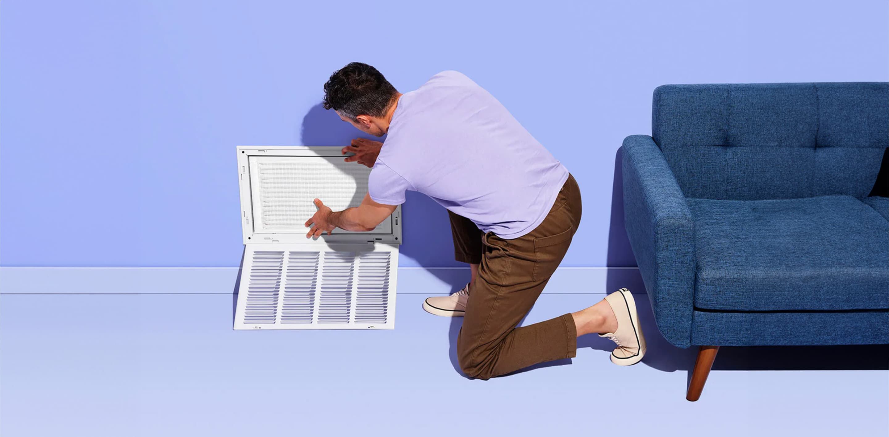 A man wearing a purple shirt is kneeling on the floor as he installs a FilterEasy Magic Filter into a wall vent.