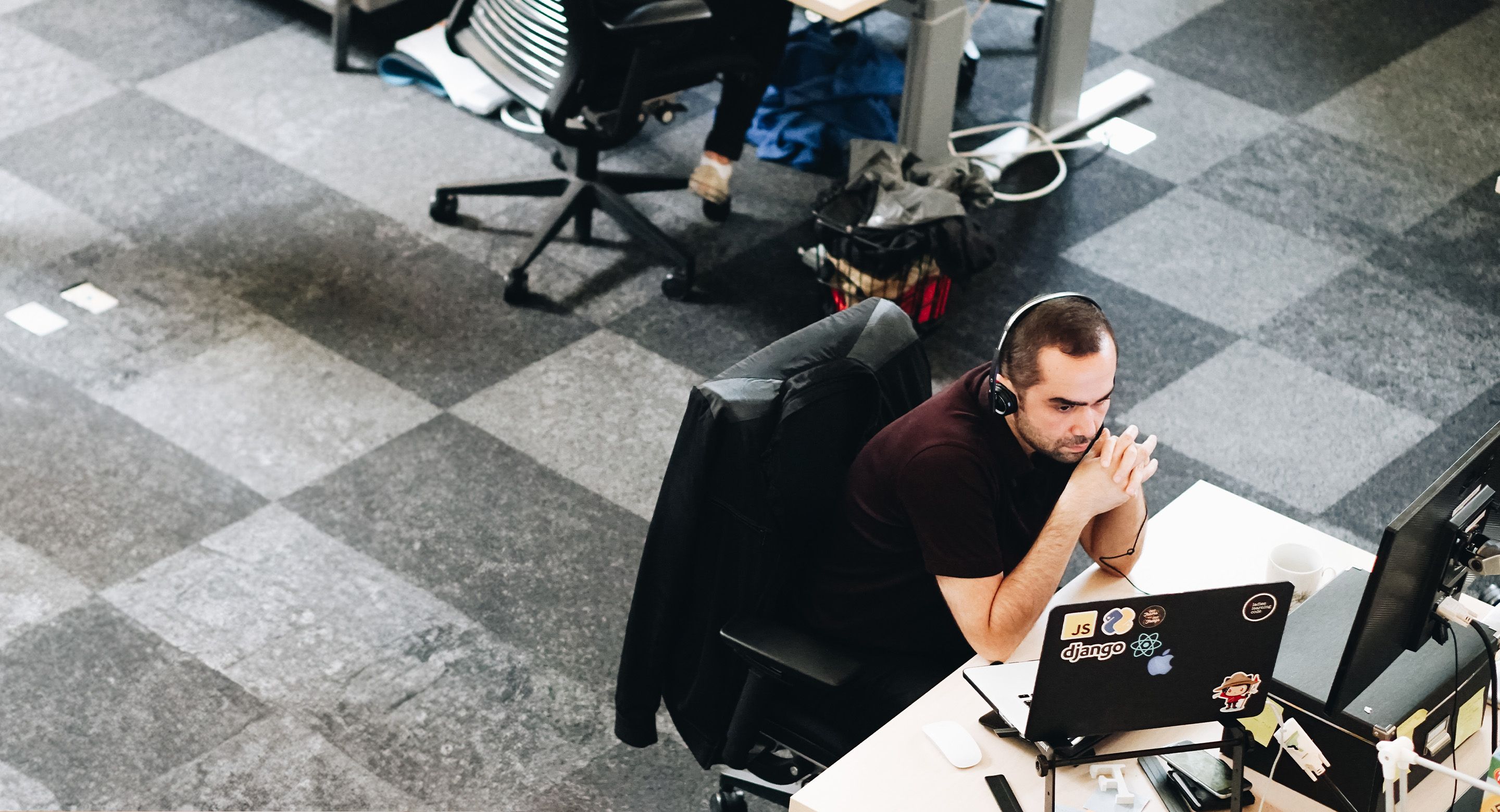 Developer wearing a headset working at a desk with a laptop covered in stickers and an external monitor in an open office environment.