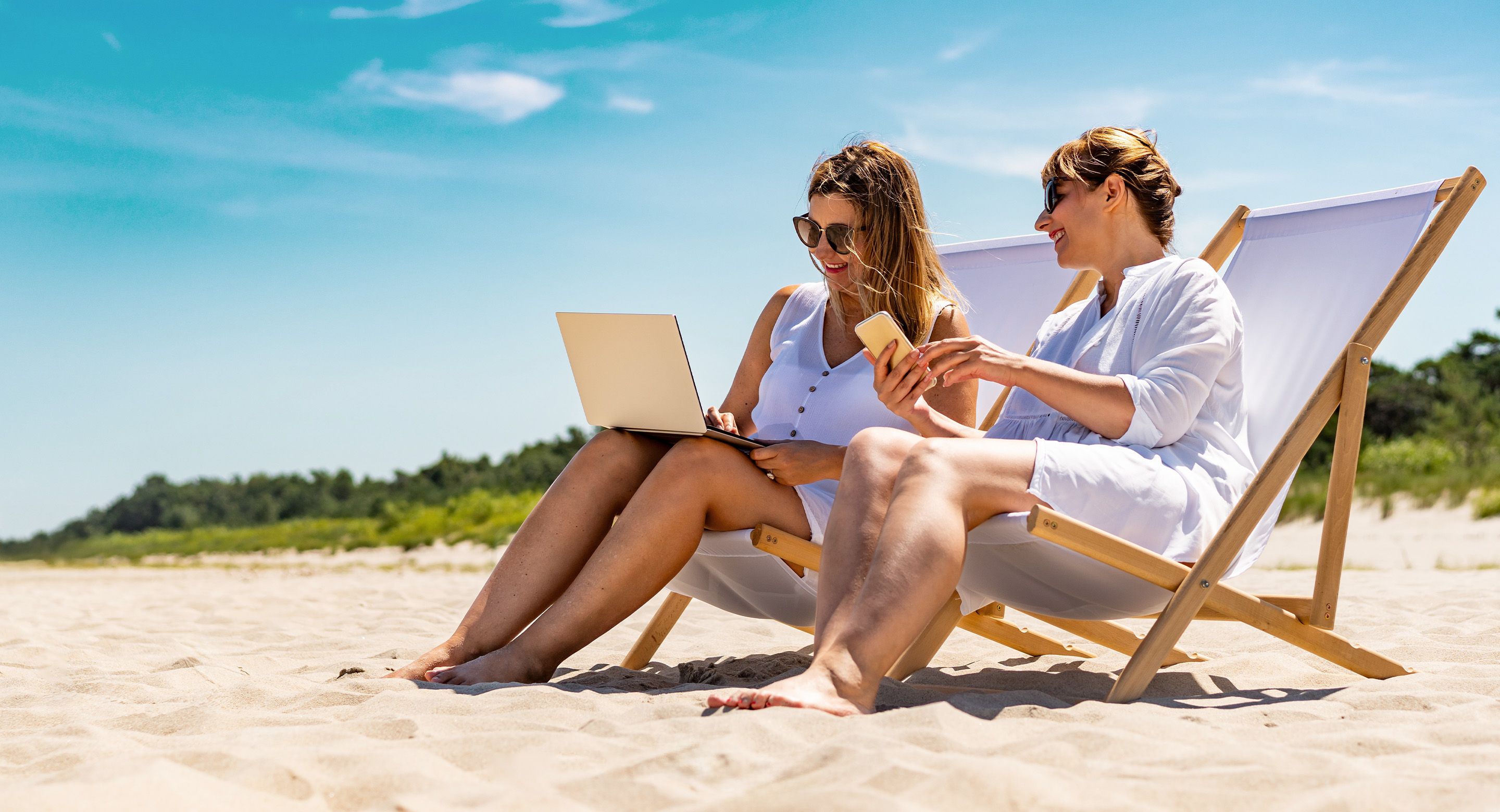 Two women sit in beach chairs on the beach with their laptop and smartphone in hand