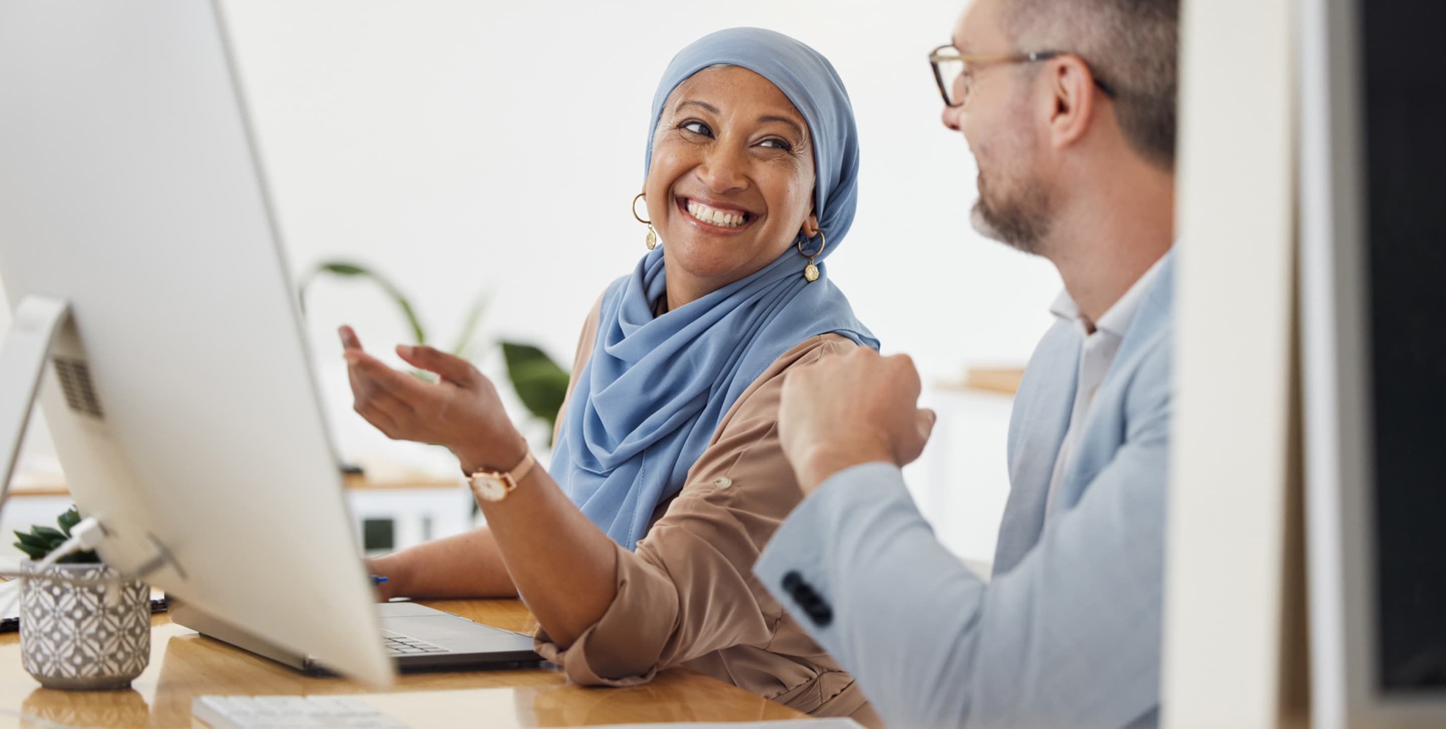 A woman looking at her coworker and smiling with one hand raised, gesturing towards a computer screen.