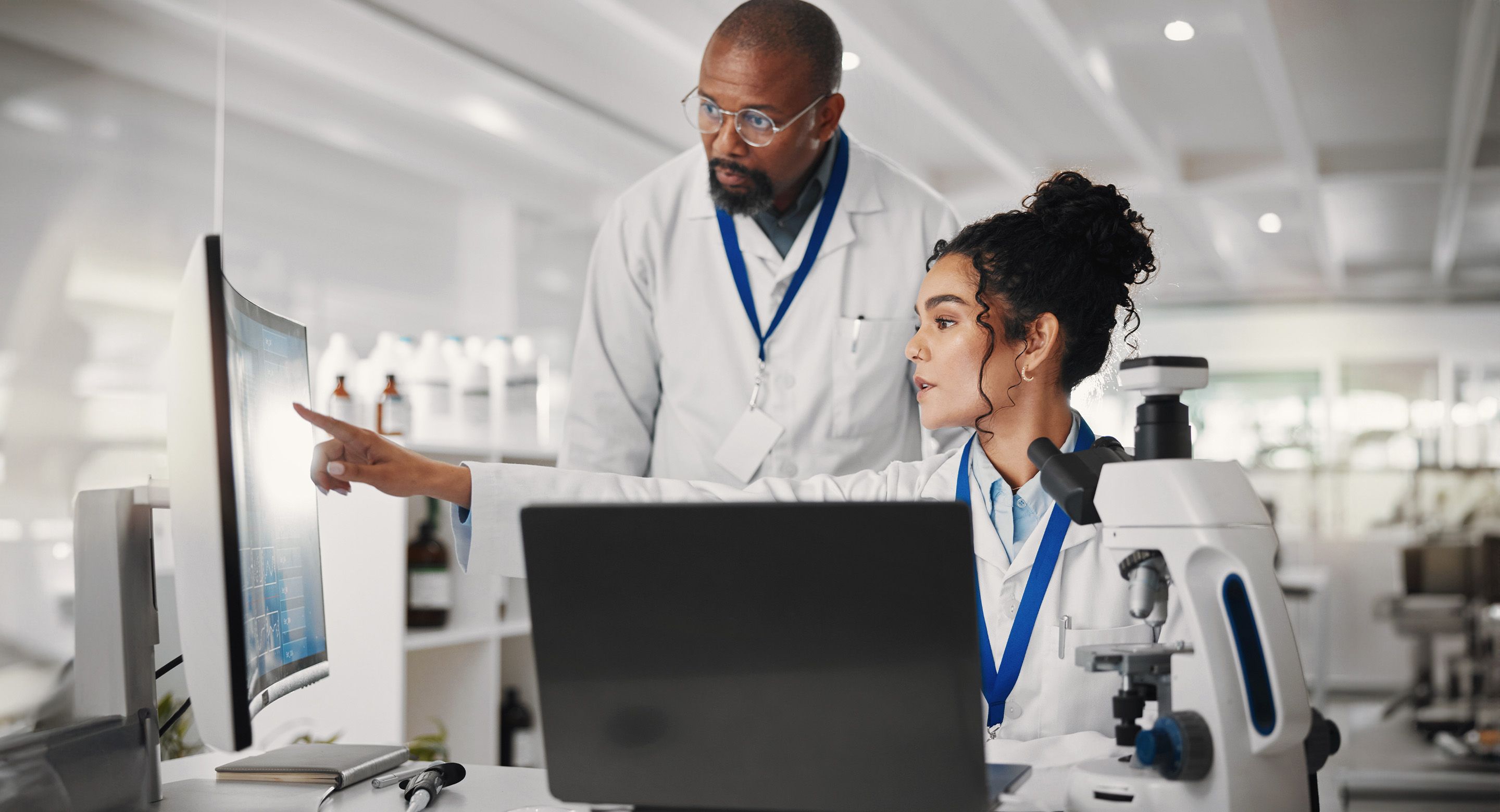 Two lab workers looking at data on a monitor beside a microscope in a laboratory.