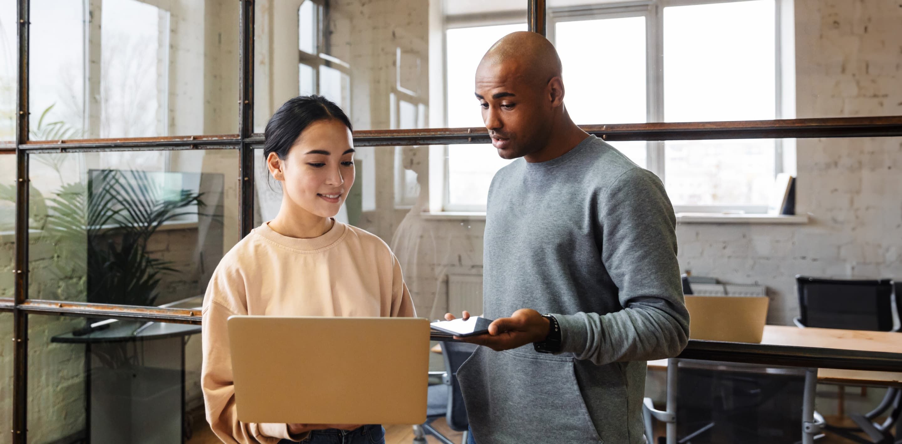 Two coworkers are in an office, standing and looking at a laptop screen.