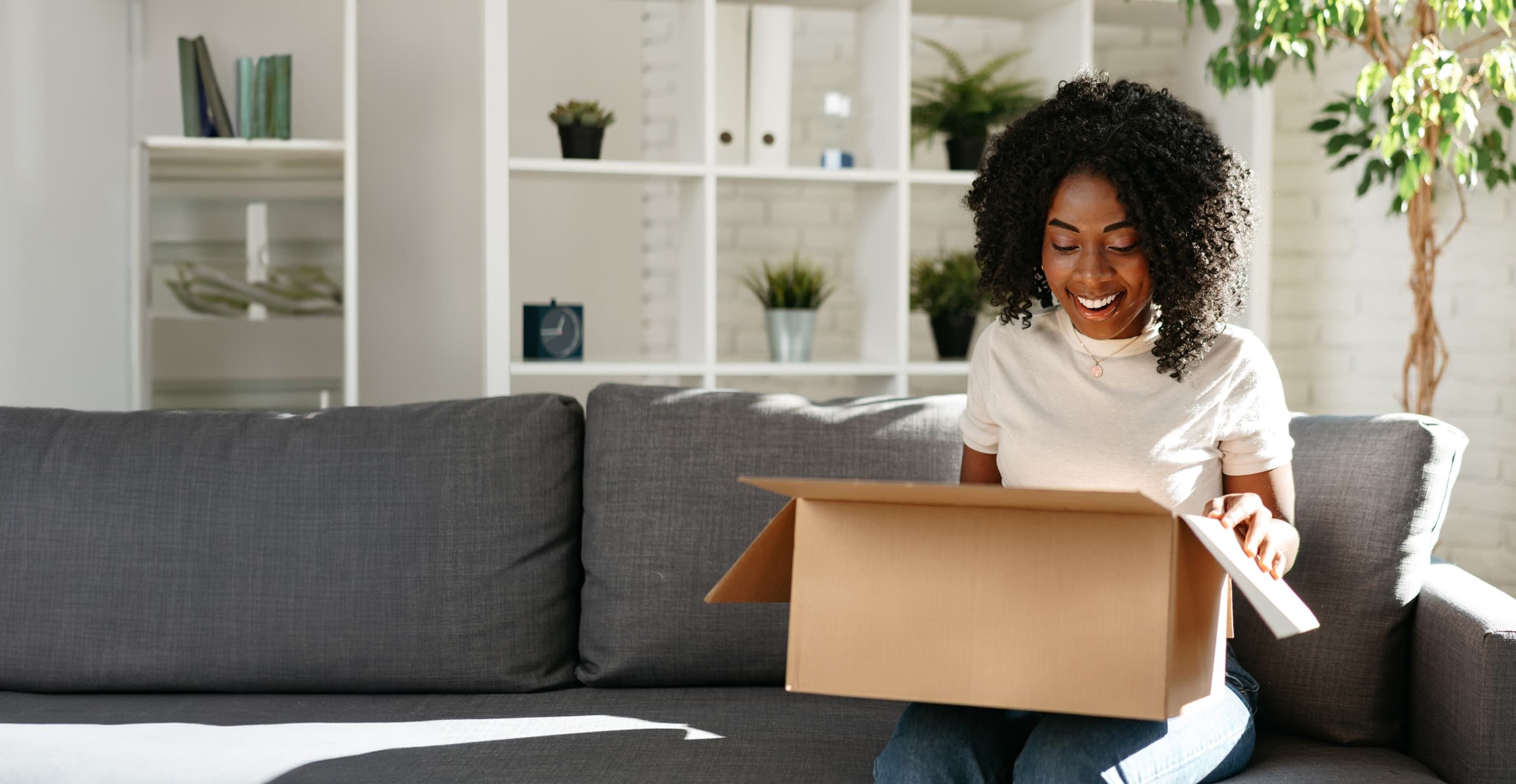 A person sitting on the end of a grey couch. They are opening a cardboard box package and smiling at the contents inside.