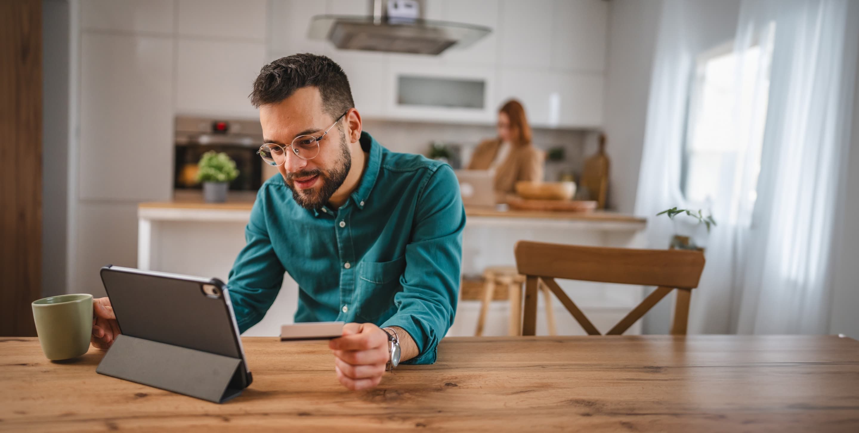 A person sitting at a kitchen table with their iPad on the surface, a green coffee mug in one hand, and a credit card in the other.