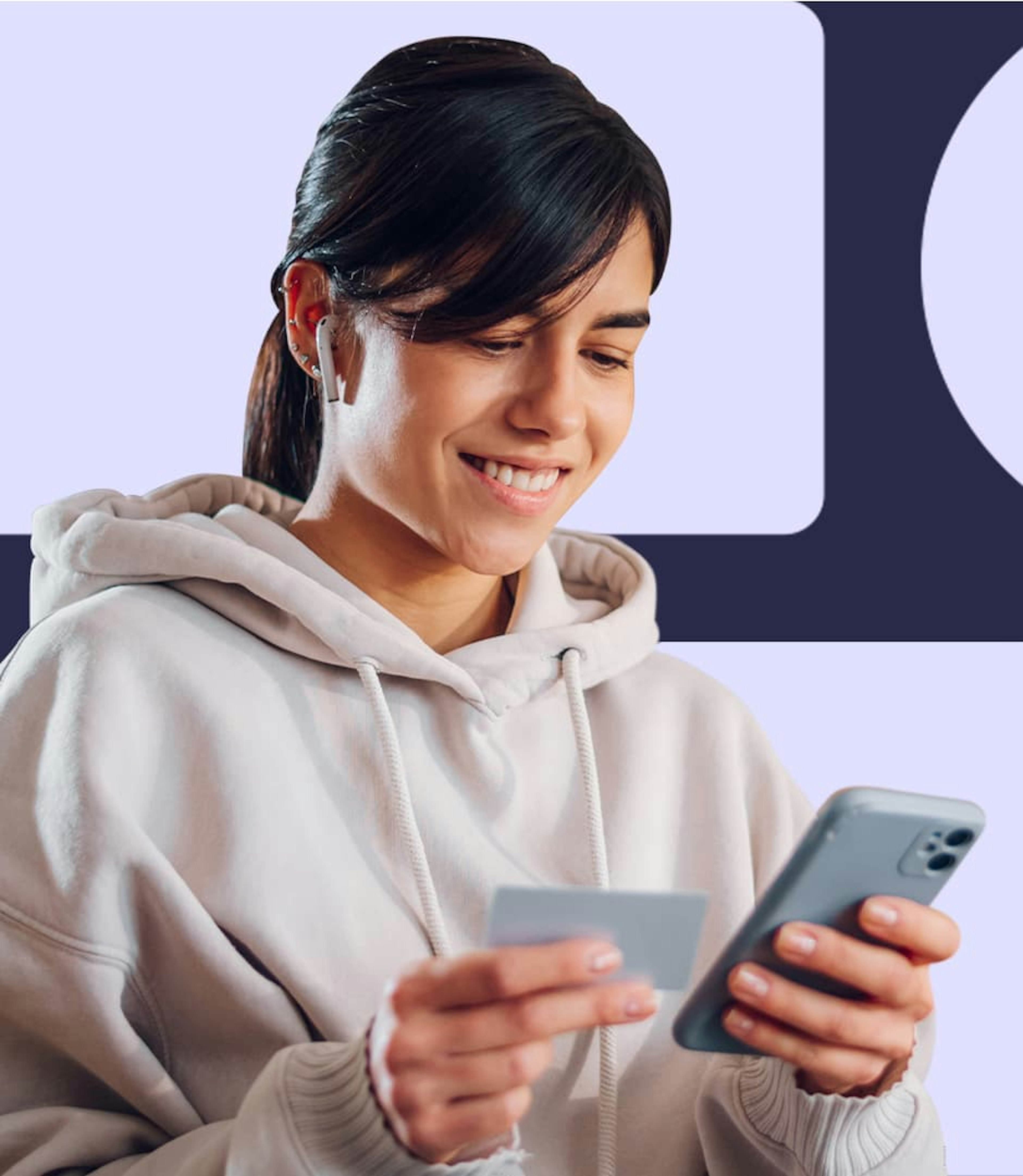 A woman wearing a beige sweatshirt, holding a payment card in one hand and a cellphone in the other.