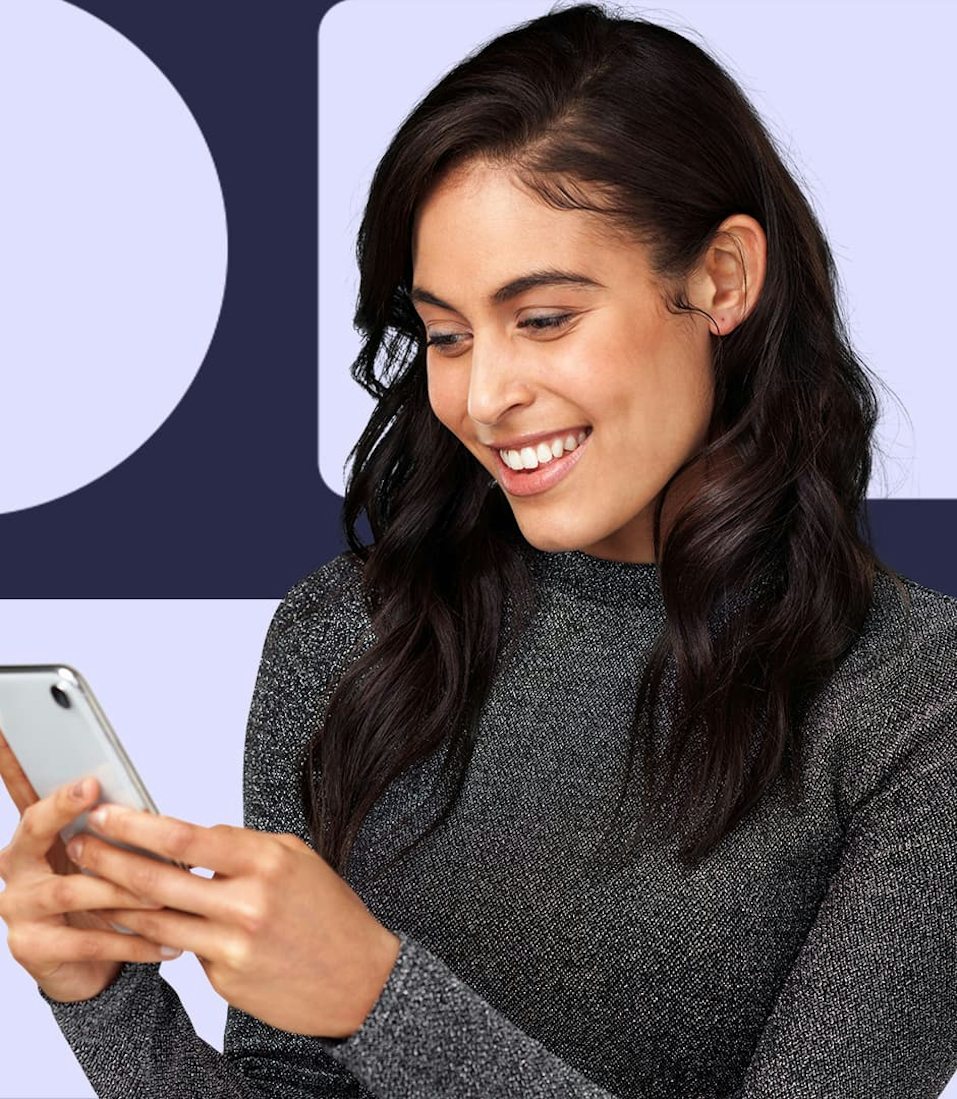 A woman wearing a grey shirt is holding a cellphone in her hands and smiling down at the screen.