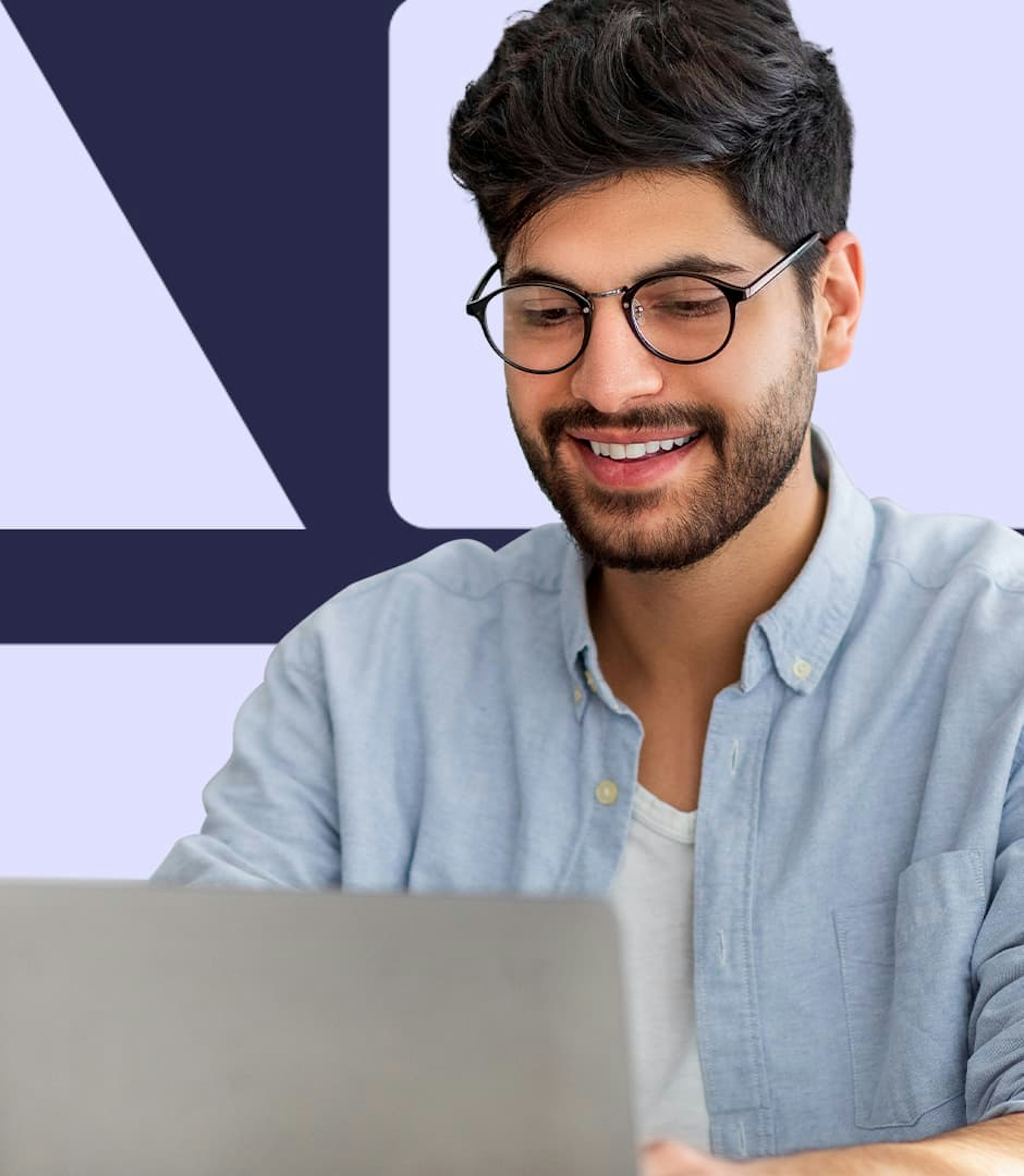 A man wearing round glasses and a casual button-down shirt is looking down at a laptop and smiling.