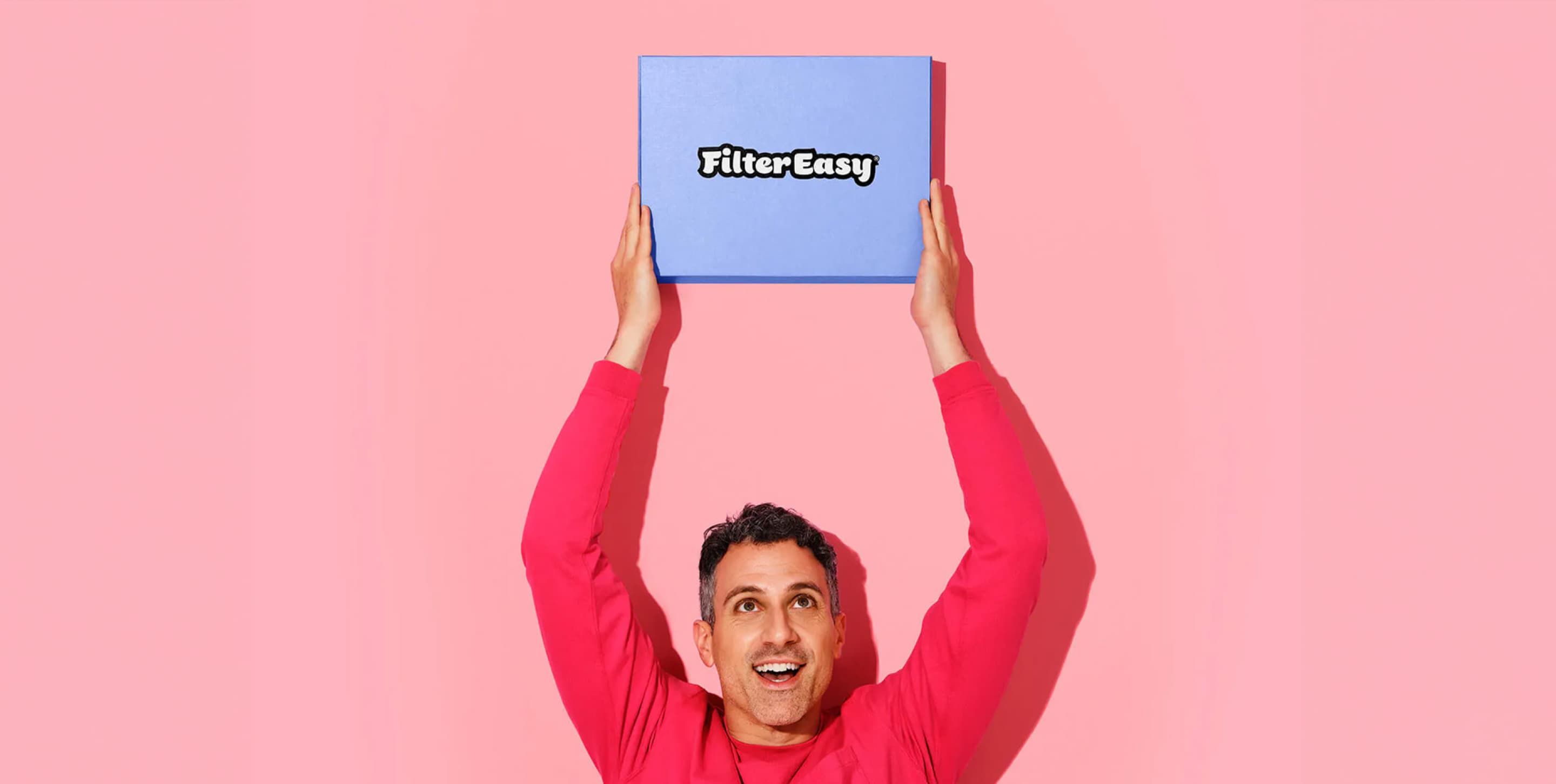 A man in a red long-sleeved shirt is smiling while holding a FilterEasy subscription box above his head.