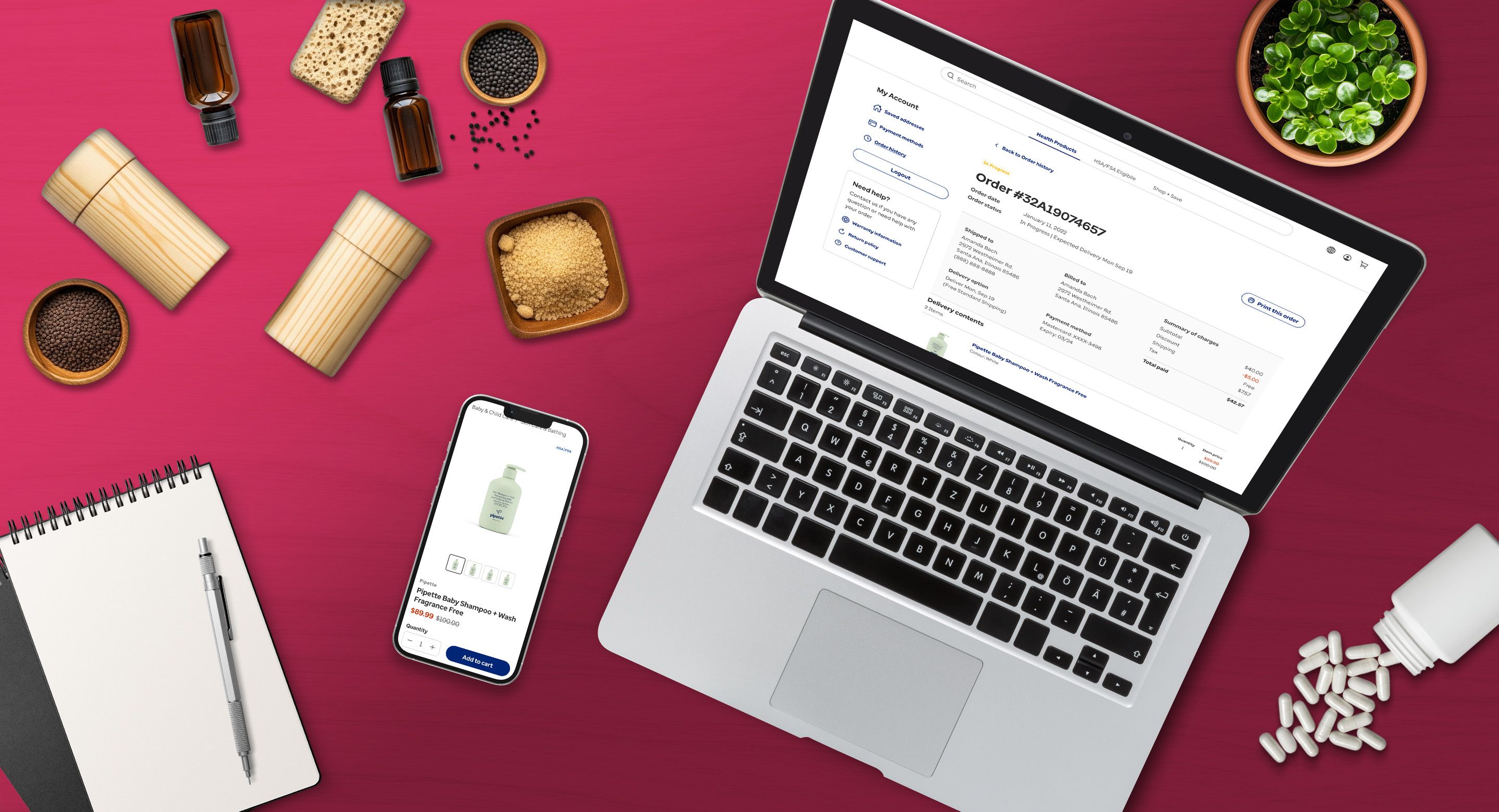 Laptop and smartphone displaying an online health and wellness store on a pink desk with natural ingredients, supplement capsules, essential oils, and a notebook.