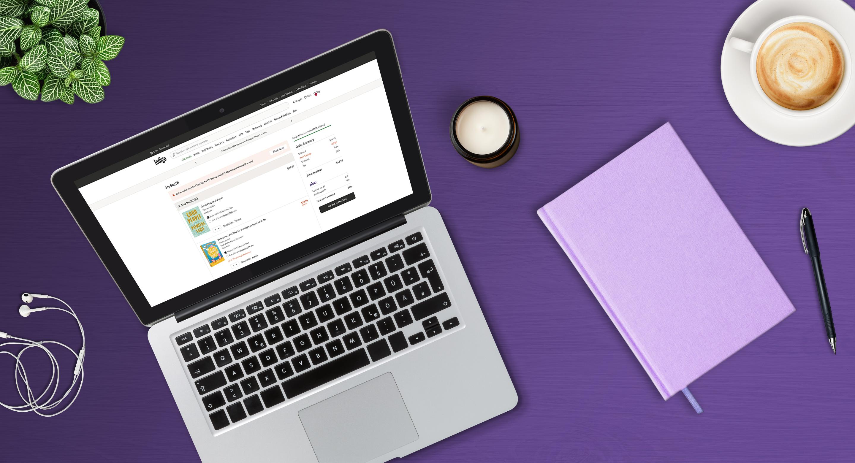Flatlay of a laptop showing the Indigo online store shopping bag with plum Rewards points, surrounded by a potted plant, a candle, a lilac notebook, a pen, and a cup of coffee on a purple desk.
