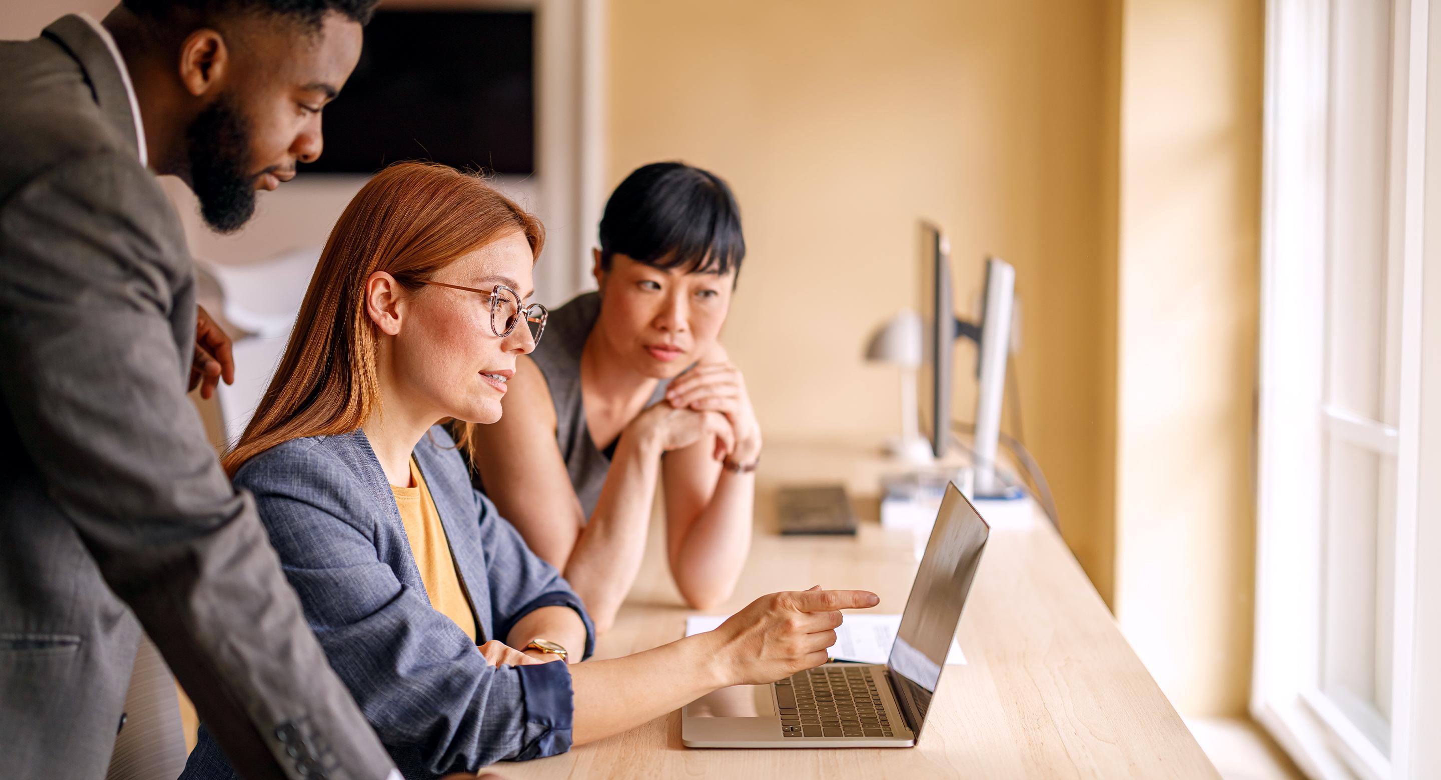 Three colleagues gathered around a laptop at a bright office window, collaborating and pointing at the screen.