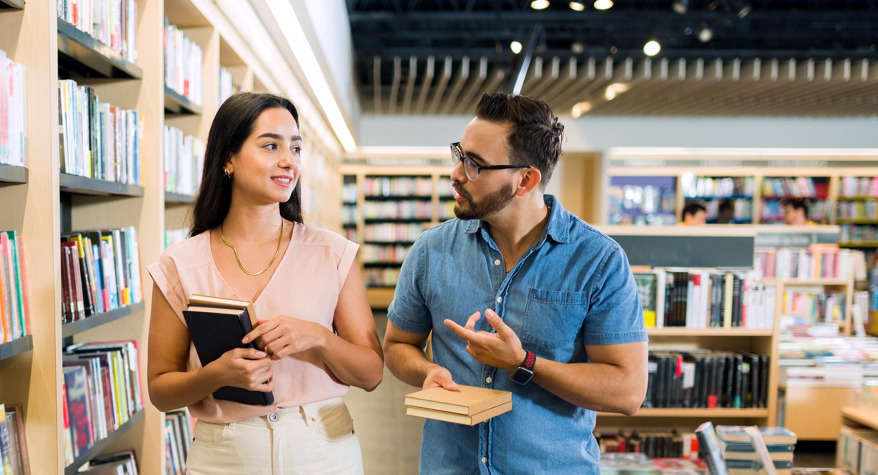 A woman and a man browsing and talking together while holding books in the aisle of an Indigo bookstore.