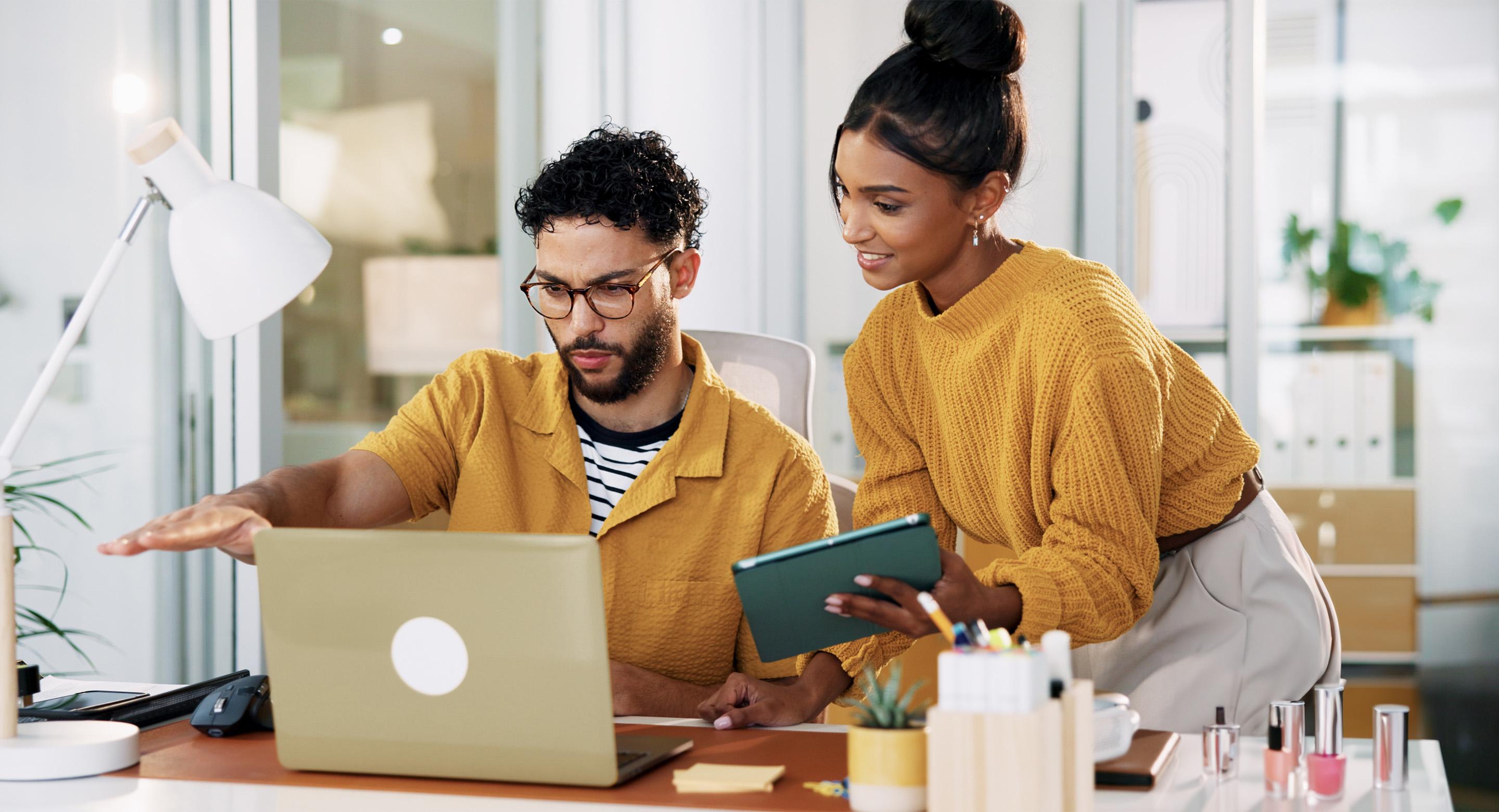 A woman looking at her coworker's screen and showing him a tablet with a beauty product on the desk.