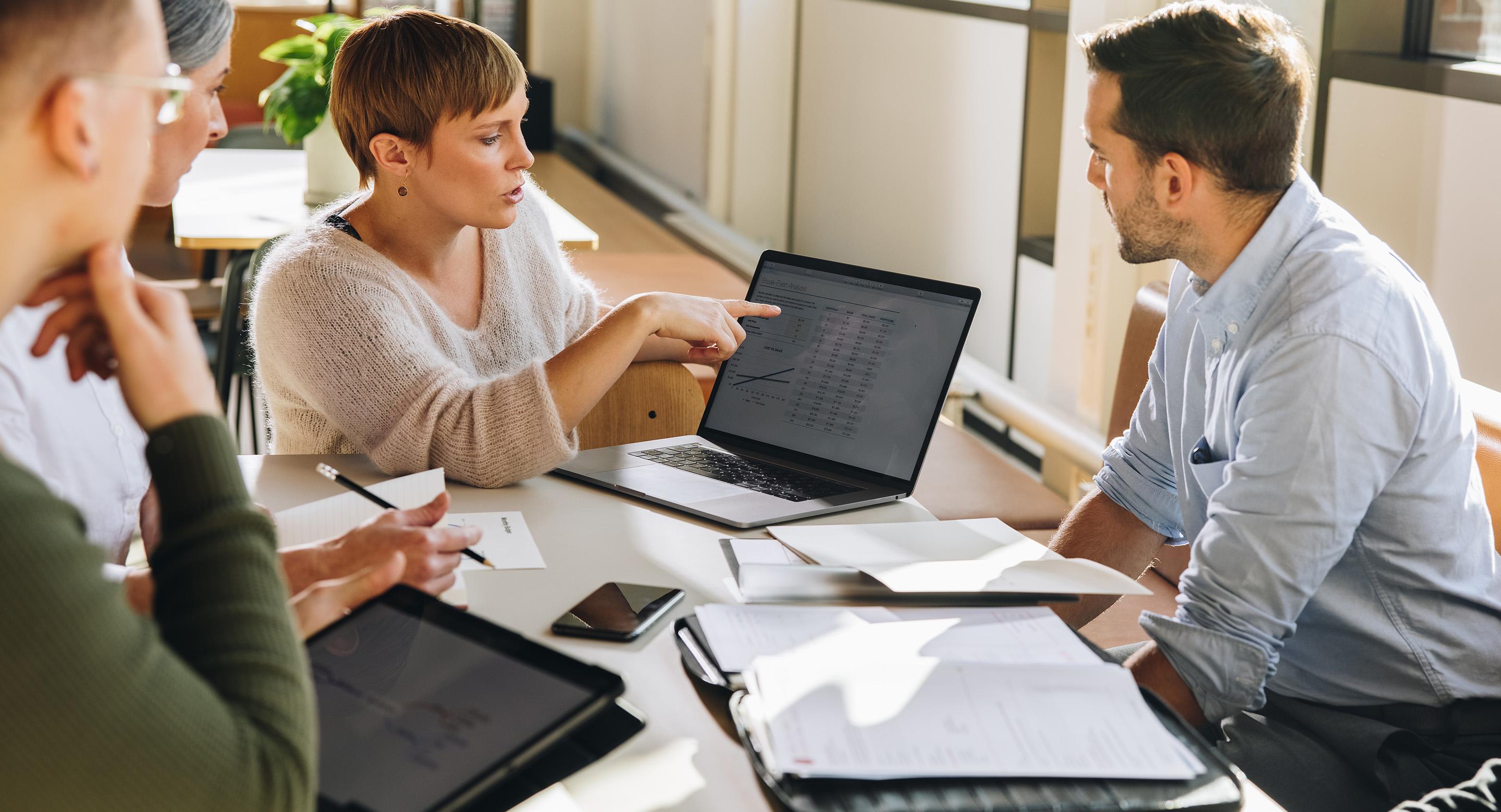 Three colleagues reviewing data on a laptop during a meeting, with documents and tablets on the table.