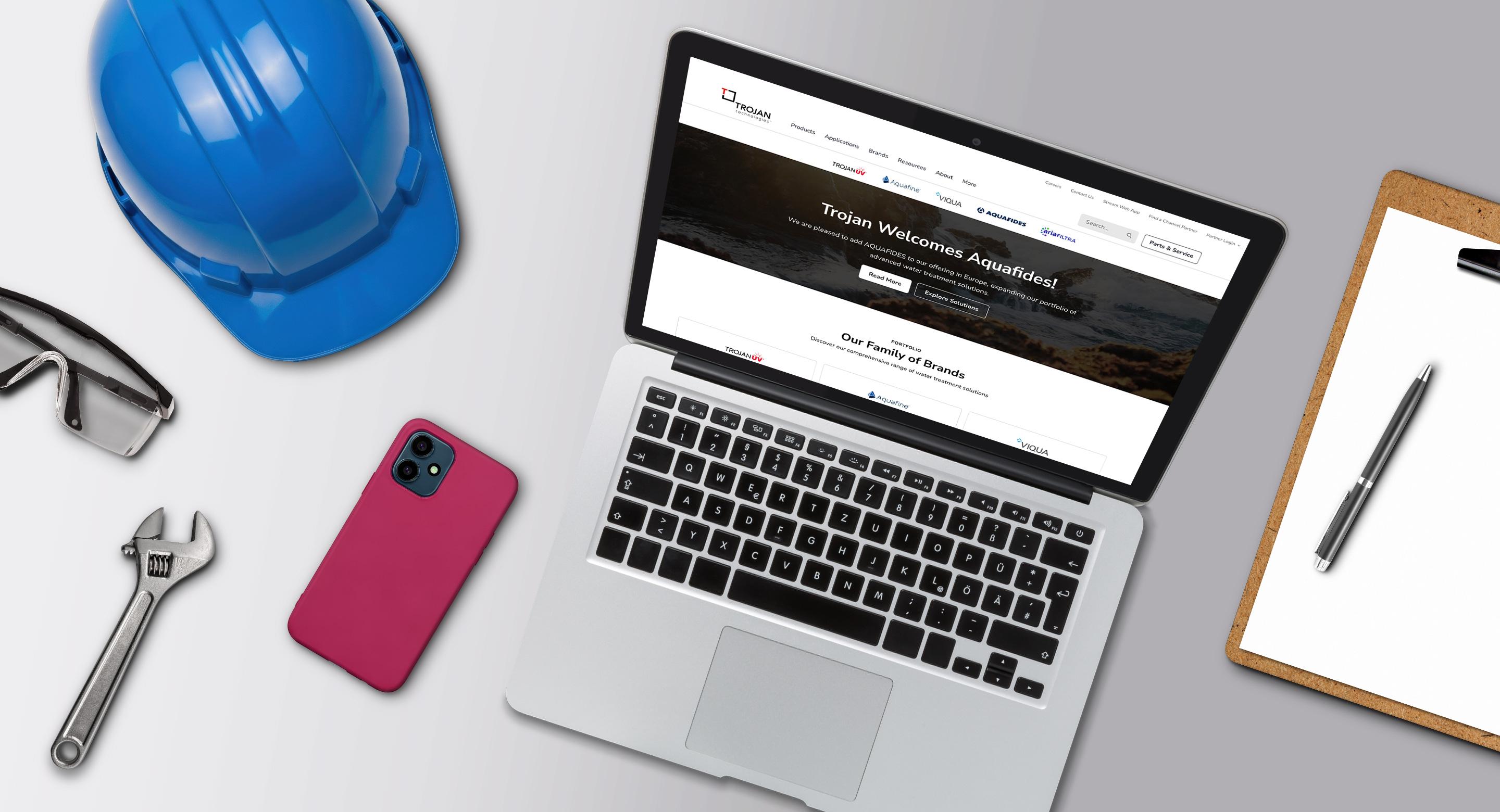Laptop displaying the Trojan Technologies website homepage, surrounded by a hard hat, safety glasses, a wrench, a phone, and a clipboard on a desk, representing water treatment fieldwork and digital tools.