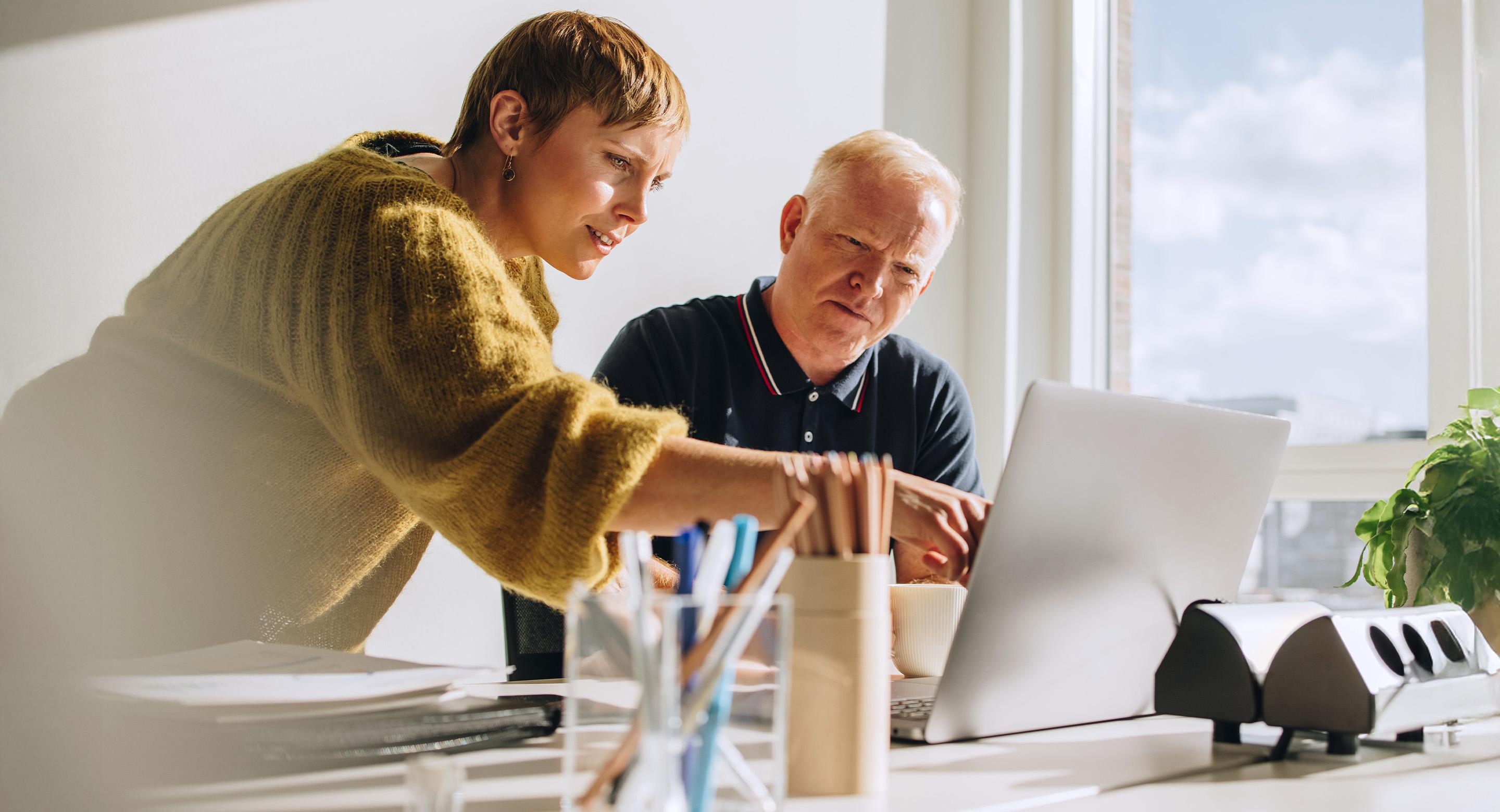 A man and woman reviewing content together on a laptop at a bright office desk.