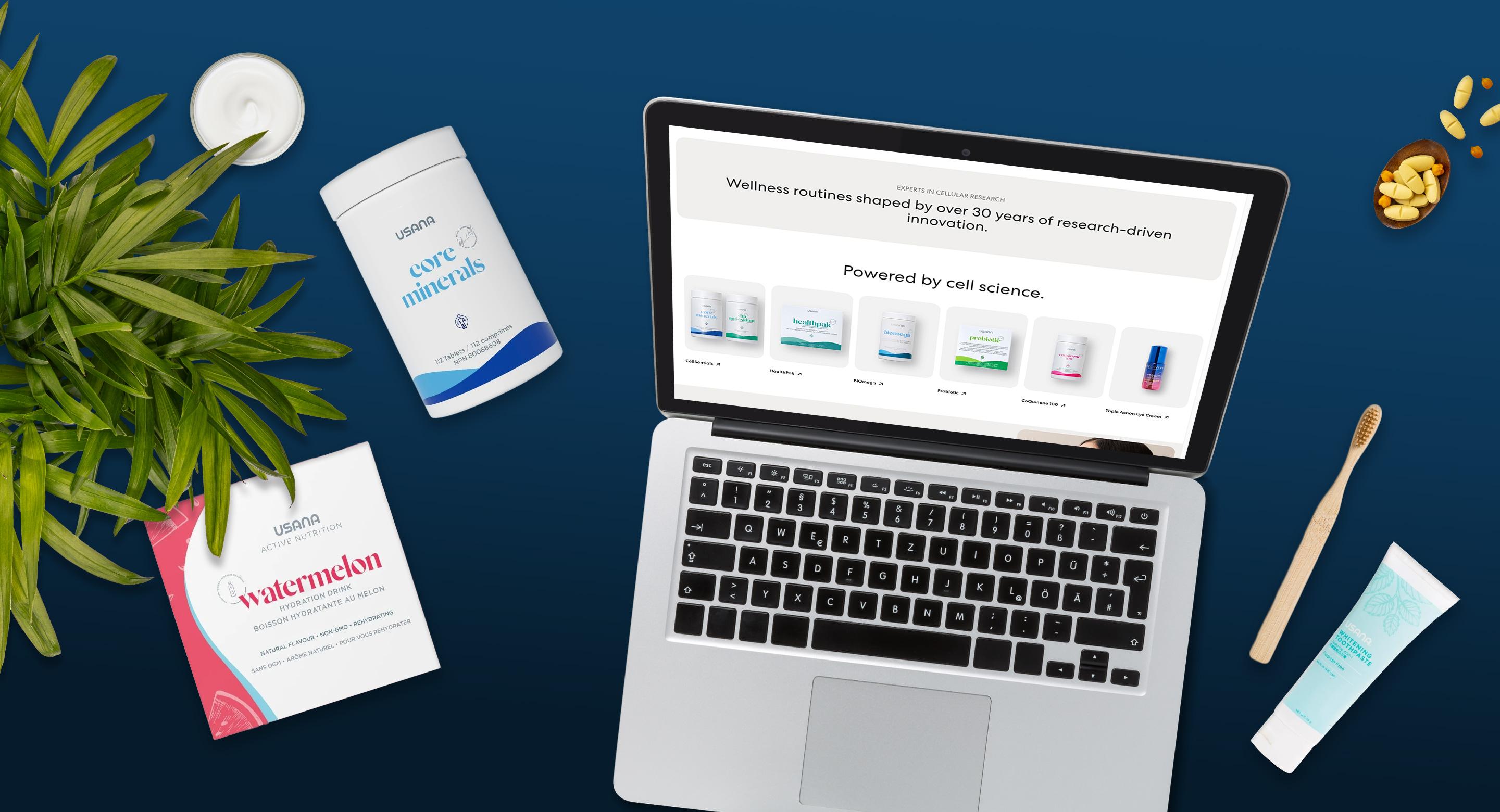 Flatlay of a laptop displaying the USANA website product grid, surrounded by USANA supplement bottles, active nutrition packets, and skincare items on a navy blue desk.
