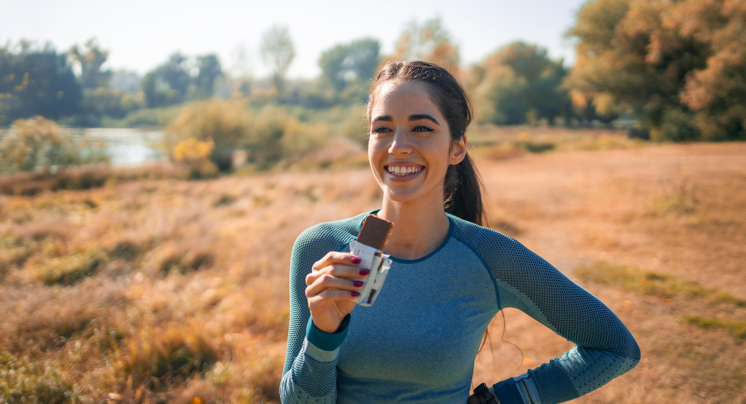 A smiling woman in athletic wear holding a USANA nutrition bar outdoors in a sunlit autumn field.