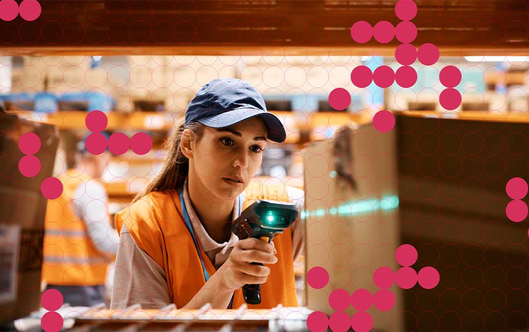 A warehouse worker in an orange vest and blue cap scans a cardboard box with a barcode scanner.