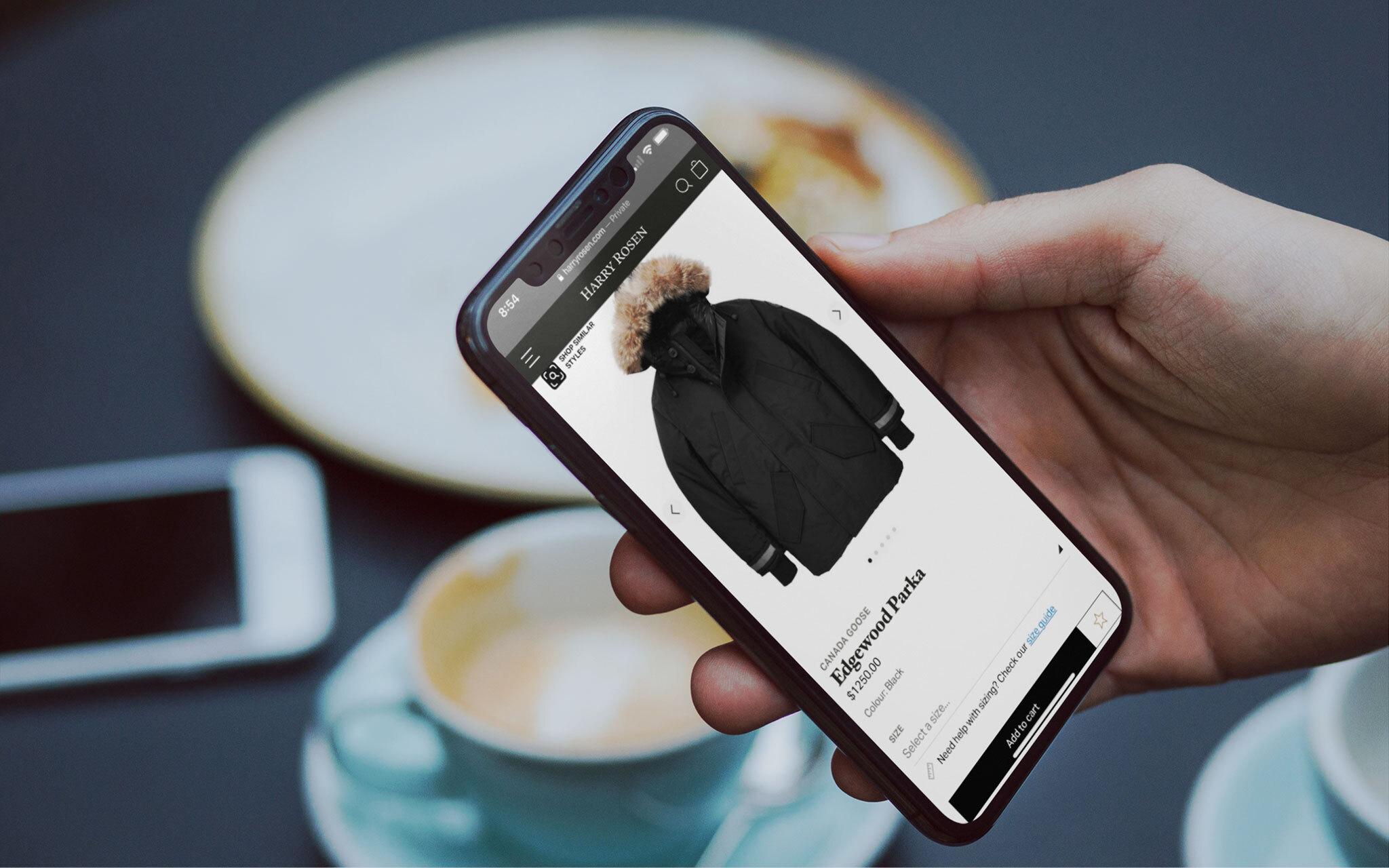 A hand holding a mobile phone browsing the Harry Rosen site. The display shows a parka that a customer can purchase online.