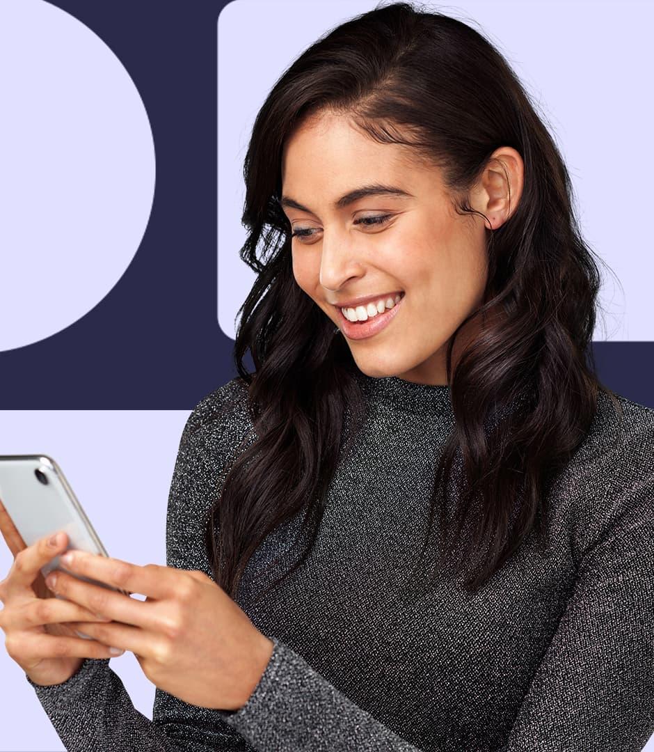 A woman wearing a grey shirt is holding a cellphone in her hands and smiling down at the screen.
