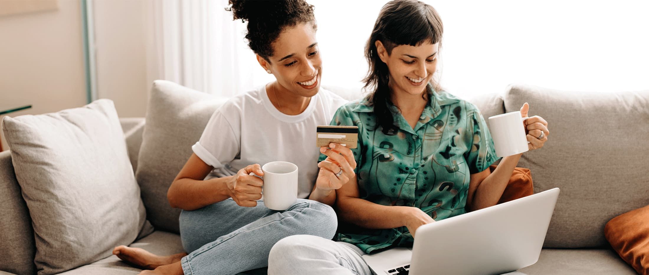 Two women sitting on a couch, one is holding a credit card in their hand, working on their laptop.