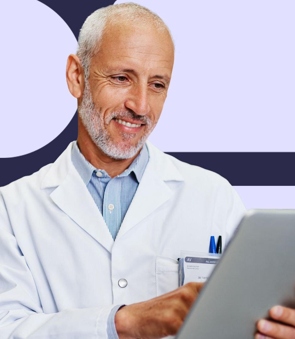 An older male doctor in a white coat, holding a tablet and smiling as he reviews information.
