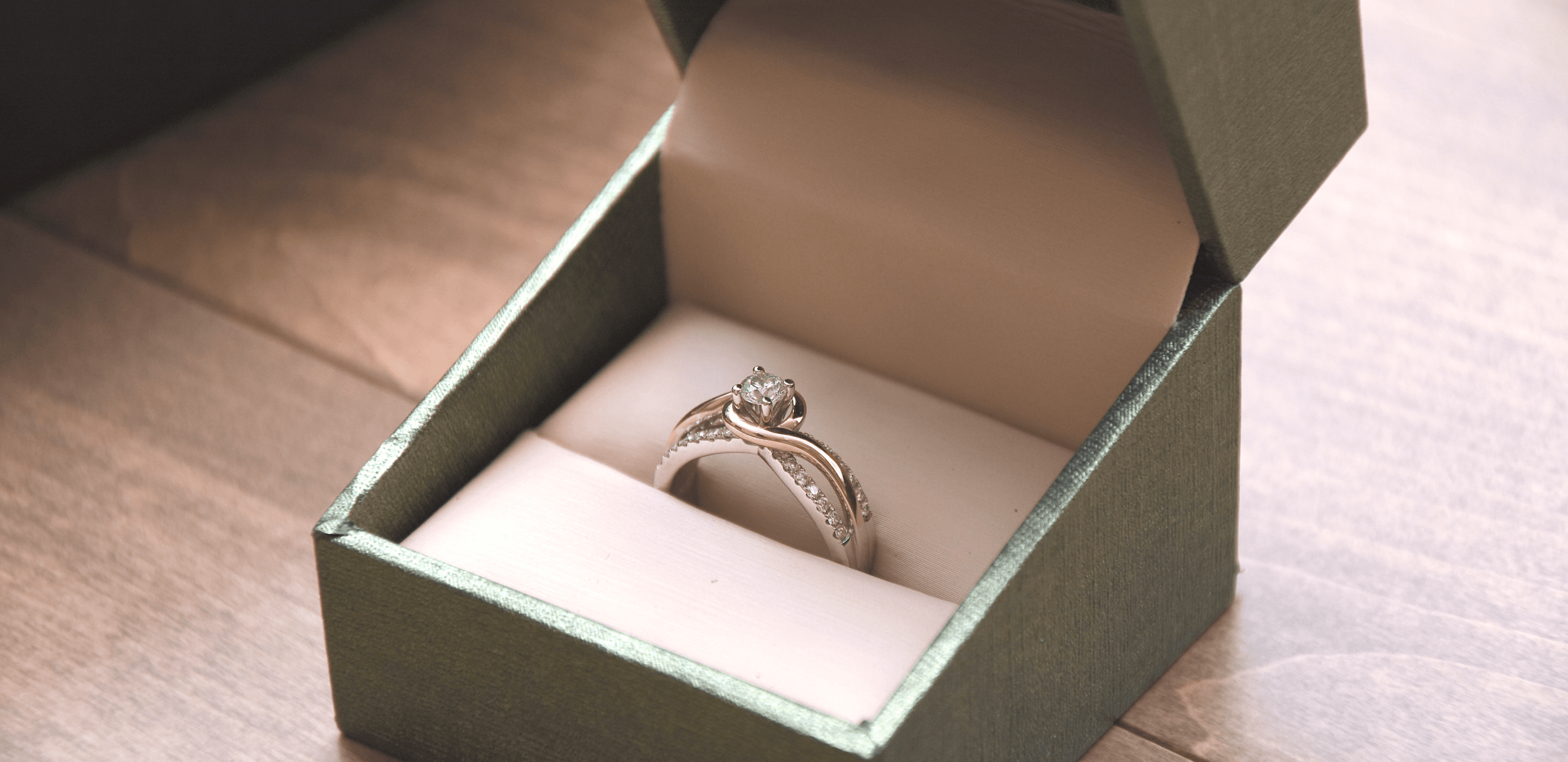 A Teilor engagement ring with an entwined band sits in a green ring box on a wooden table.