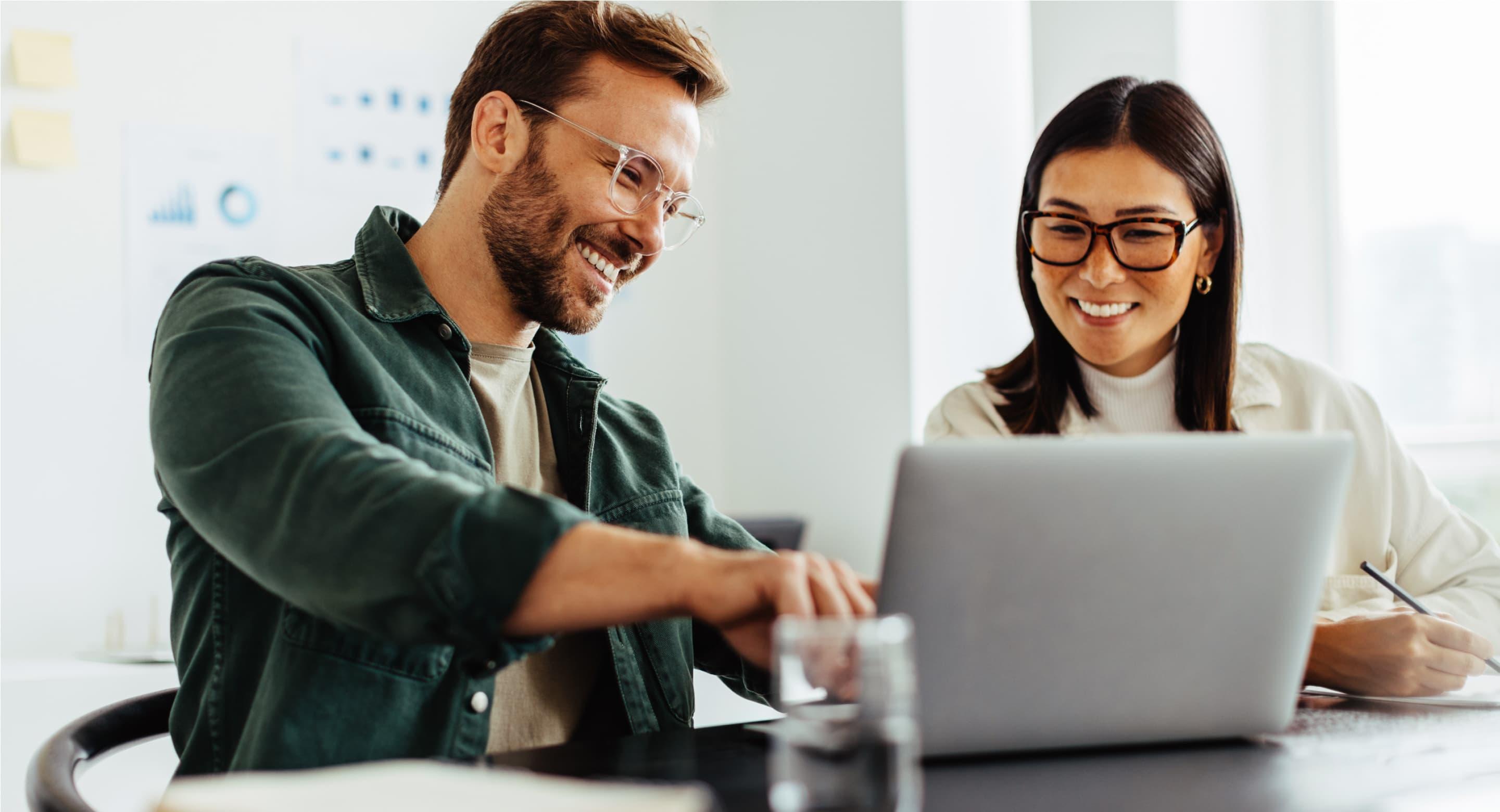 Two colleagues sitting at a table, pointing and smiling at a laptop screen.