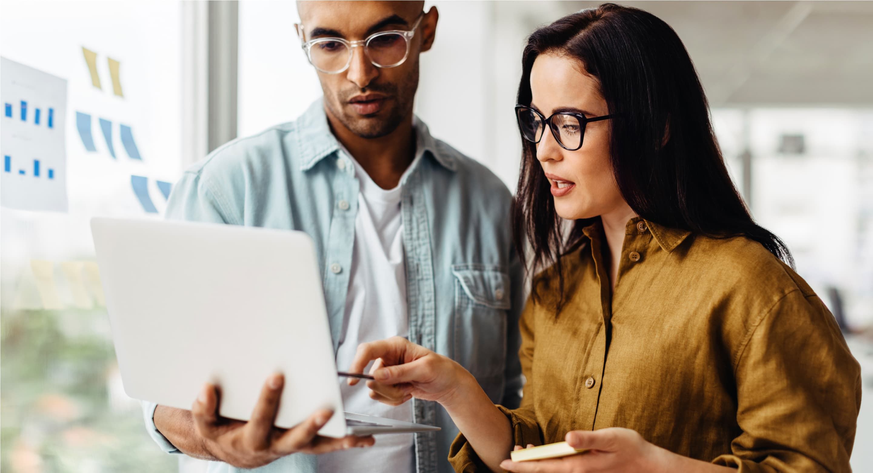 Two colleagues standing up and looking at a laptop screen. One is holding the laptop and the other is pointing to the display.