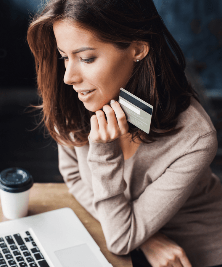 A woman with a credit card sits at a table with her laptop, coffee cup, and cellphone.