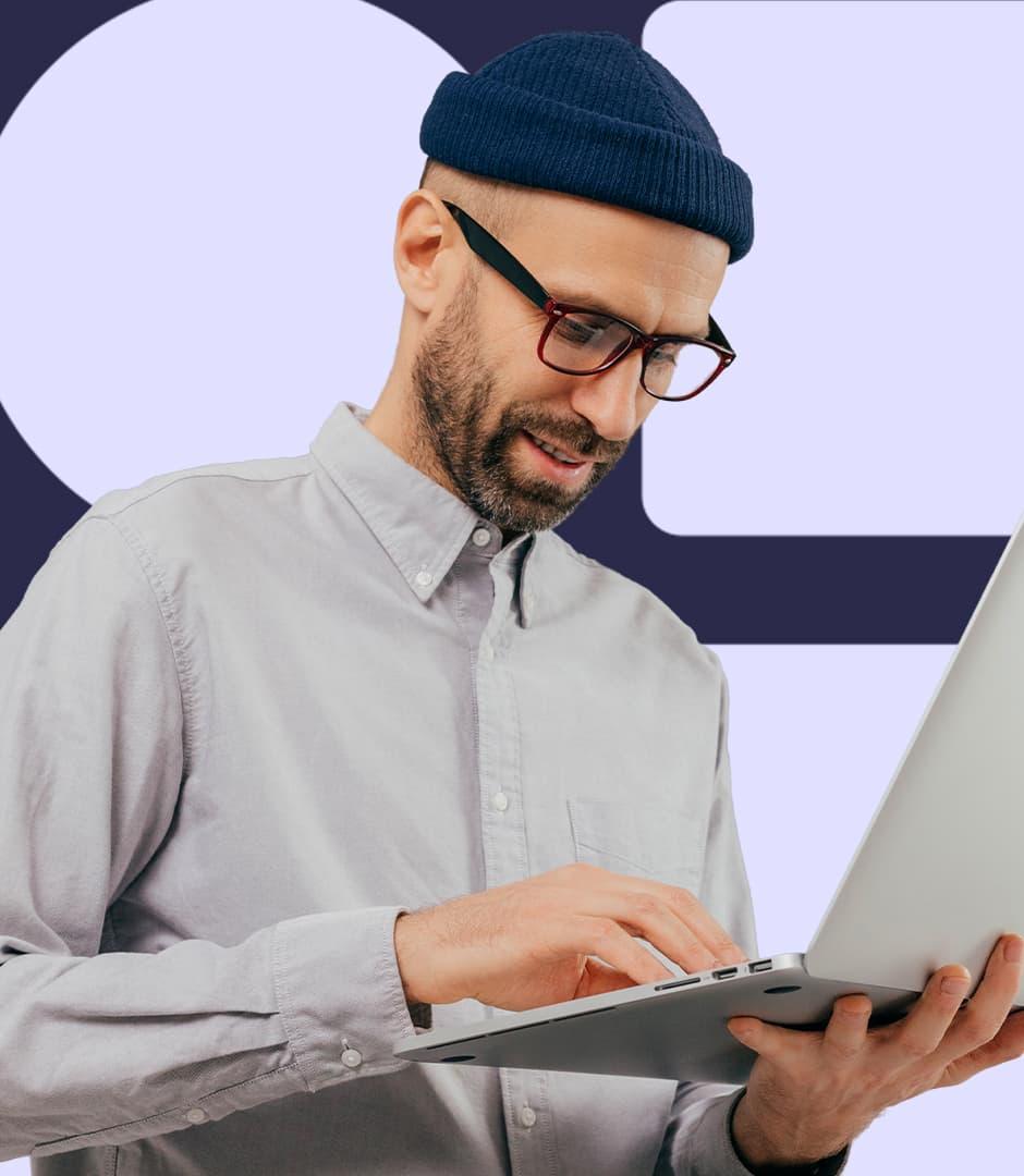 A man wearing a grey shirt and a navy beanie is holding a laptop and working.