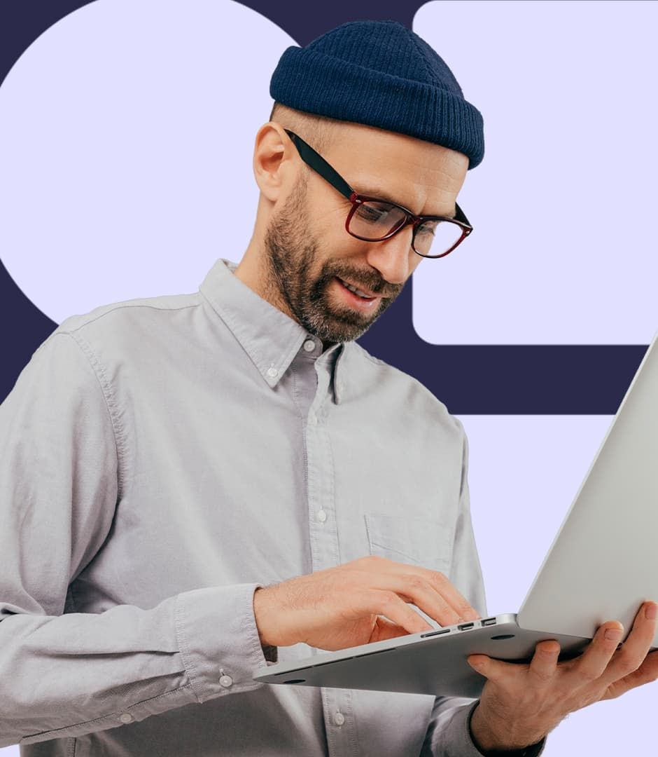 A man wearing a grey shirt and a navy beanie is holding a laptop and working.
