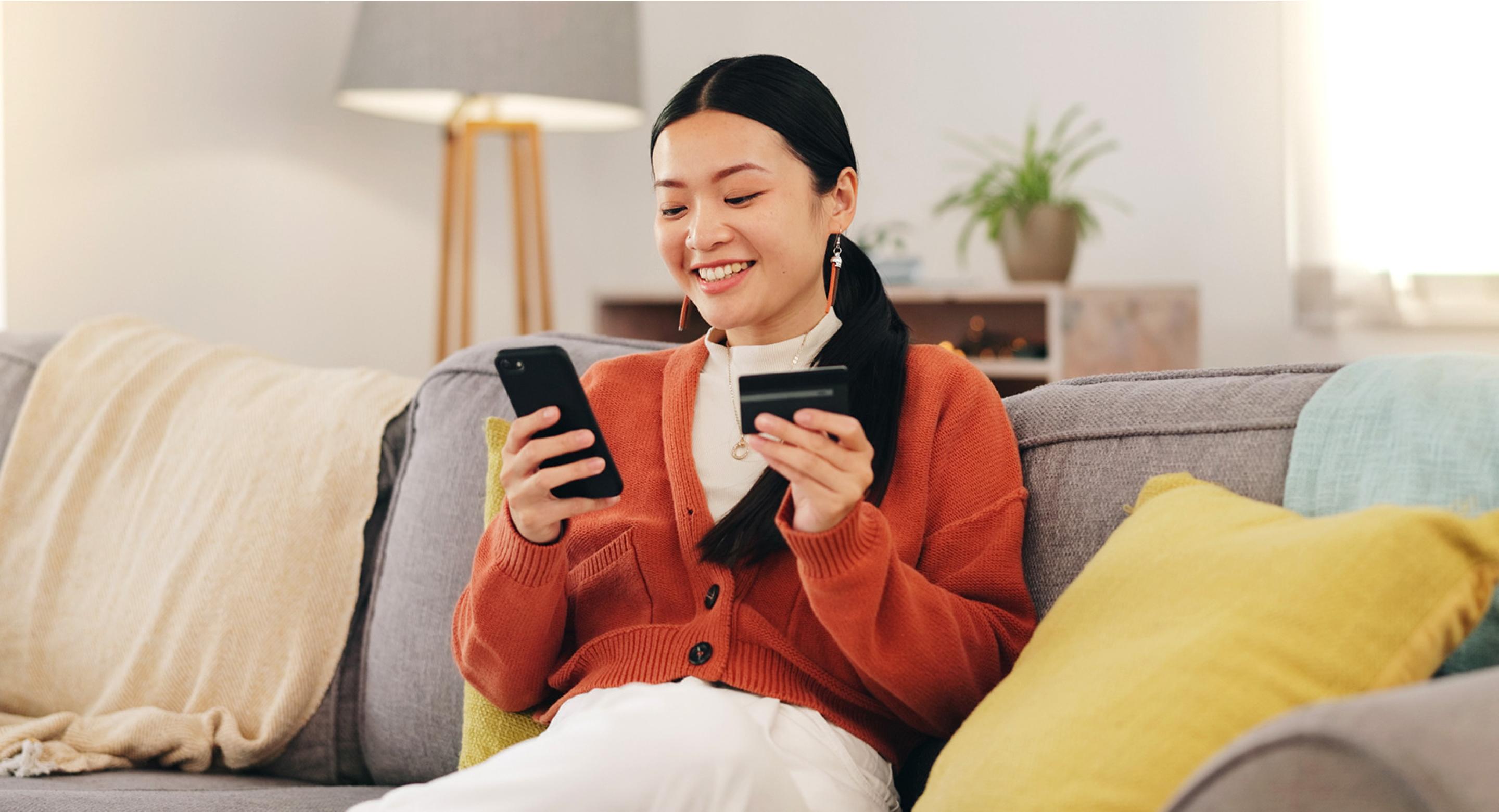 A photo of a woman sitting on a sofa with a smartphone in one hand and a credit card in the other. She appears to be shopping online using her mobile phone in the comfort of her own home.