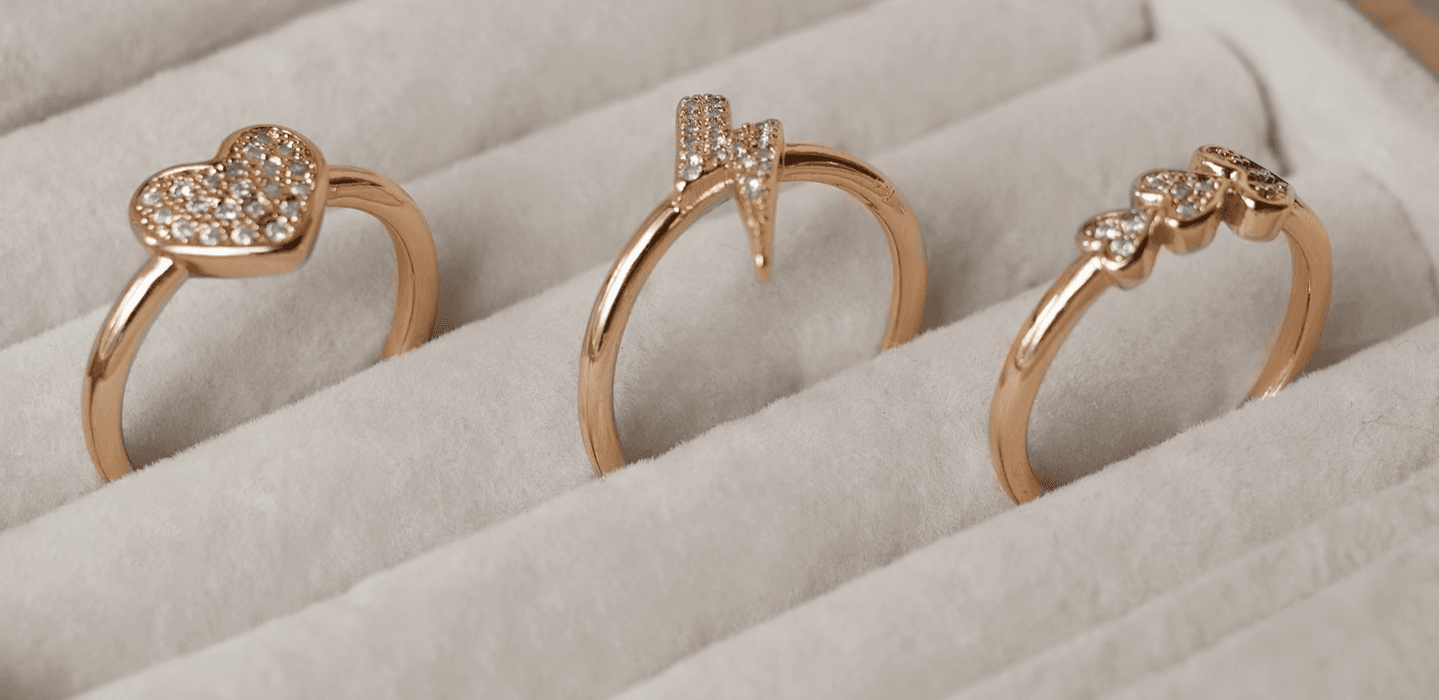Three gold Teilor rings are in a jewellery box. One has a heart, one has a lightning bolt, another has a trio of hearts.